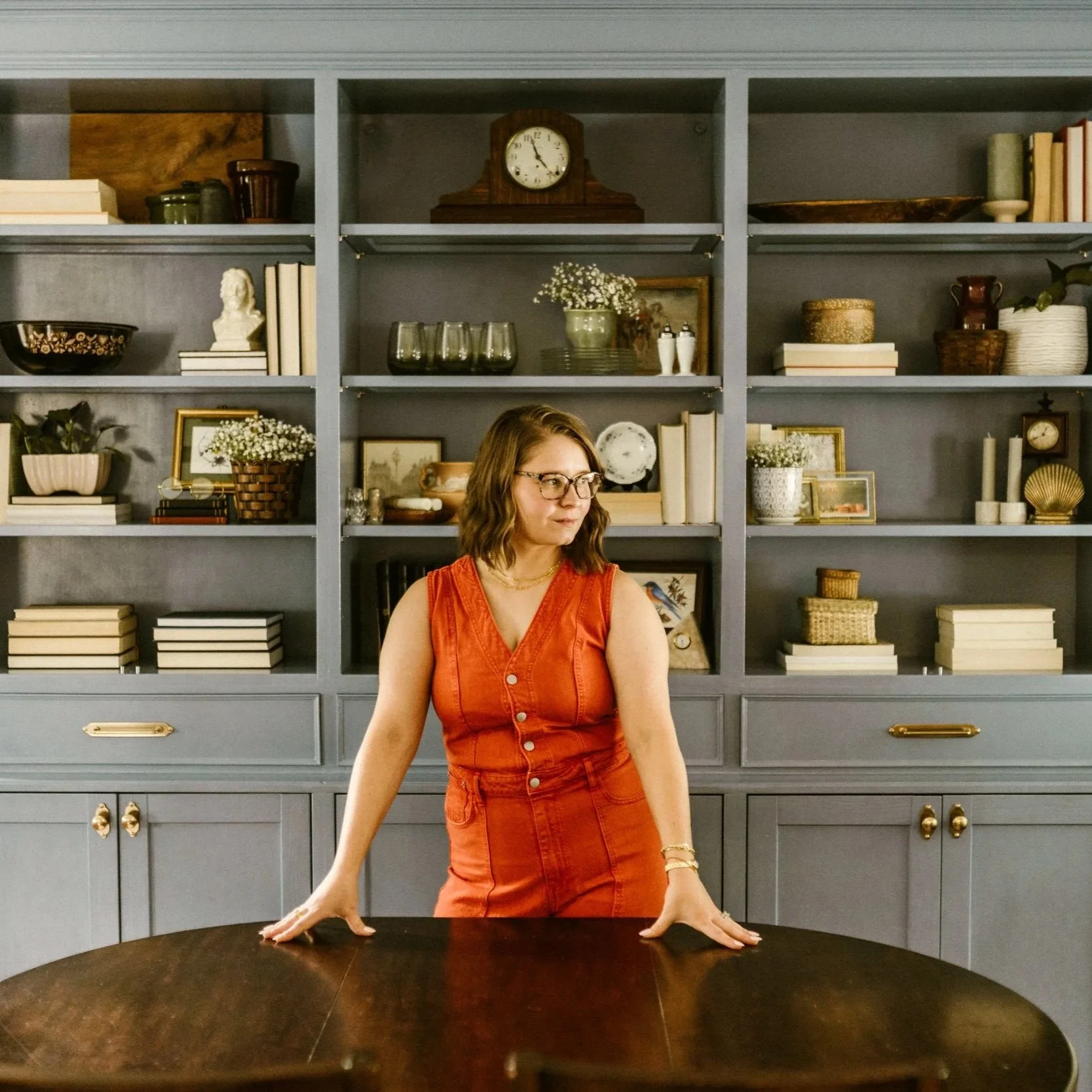 A woman in a sleeveless red dress with glasses and wavy shoulder-length hair leaning on a dark wooden table in front of a blue shelving unit filled with books, decorative items, and plants.