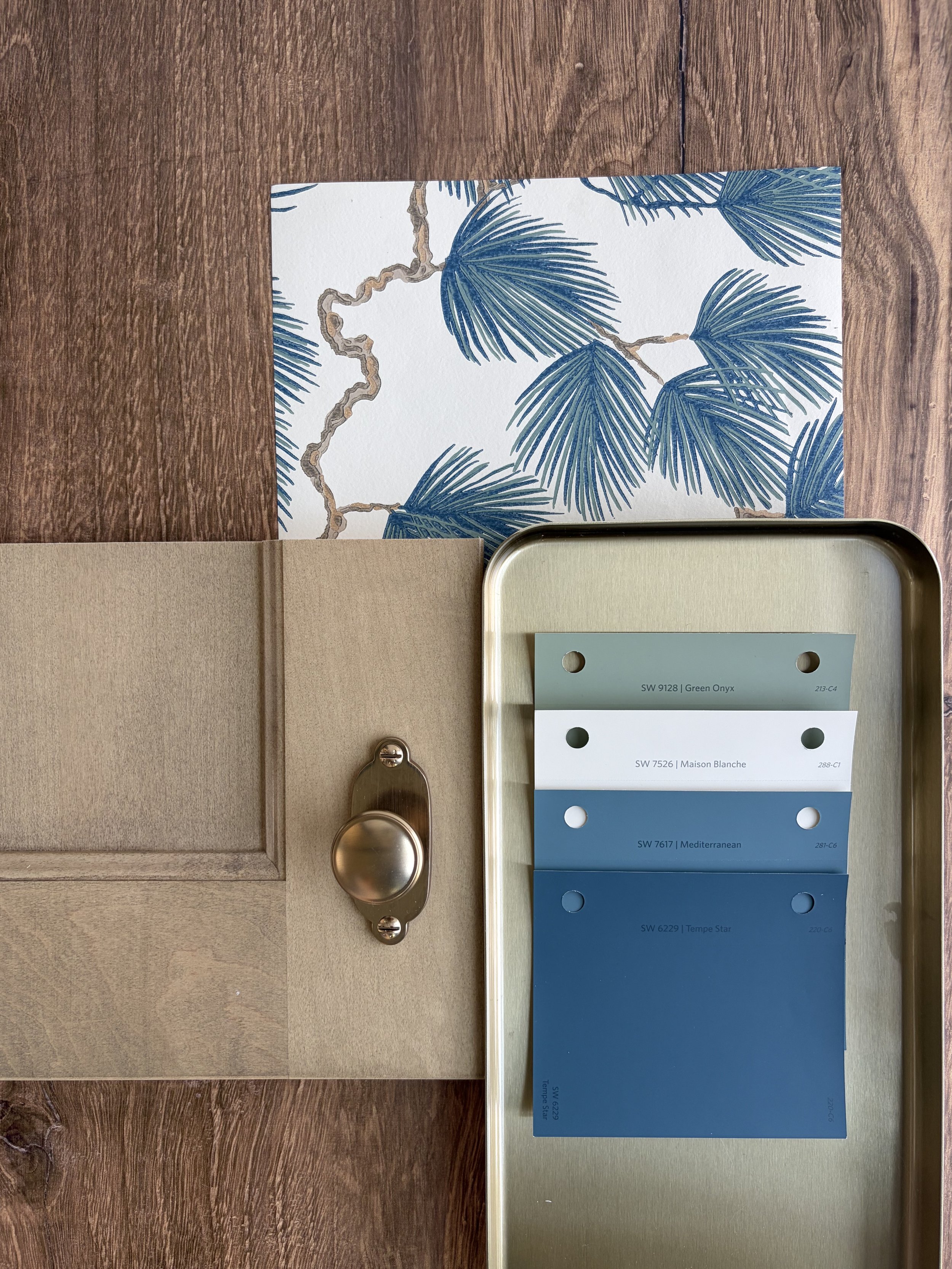 Color palette sample book with various shades of blue, green, and white, placed on a beige tray with a gold handle, and a decorative card with blue palm leaves behind it.
