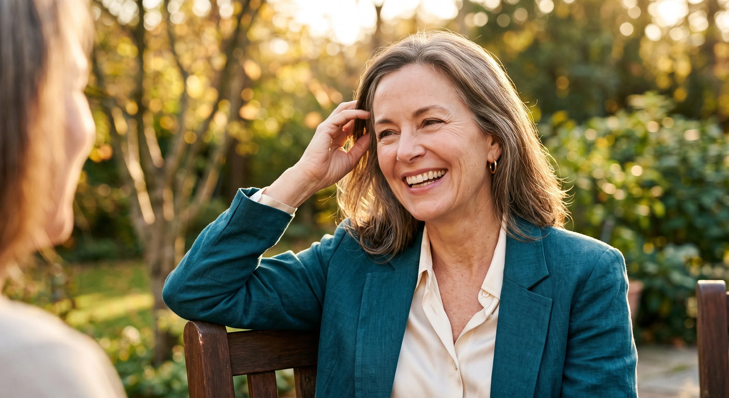 A mature woman with natural grey hair tucking her hair behind her ear outdoors in warm golden-hour light, healthy skin and a soft smile.