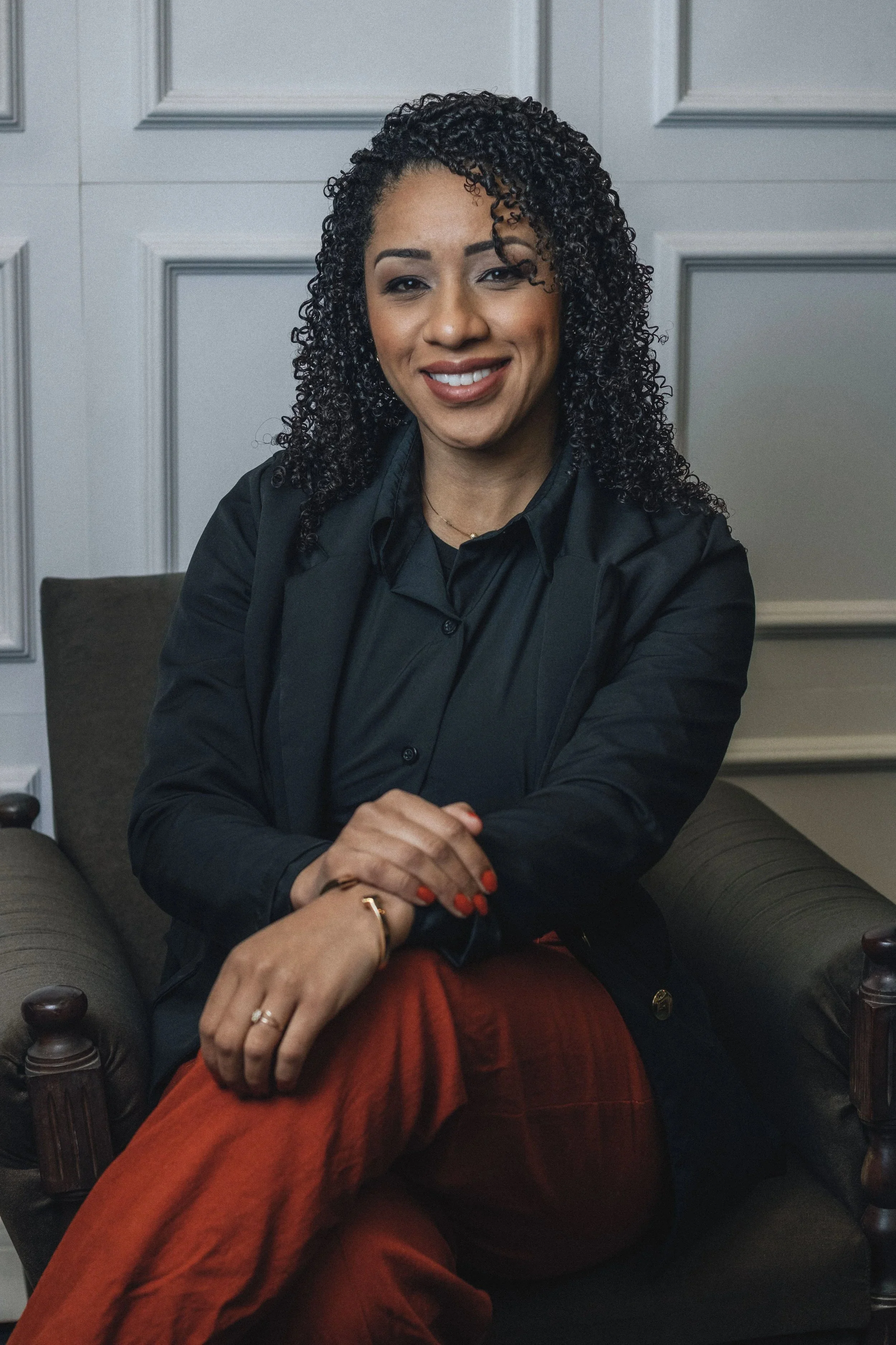 A woman leader physician with curly hair in a black blazer and red pants sits in an armchair, smiling, hands clasped in her lap.