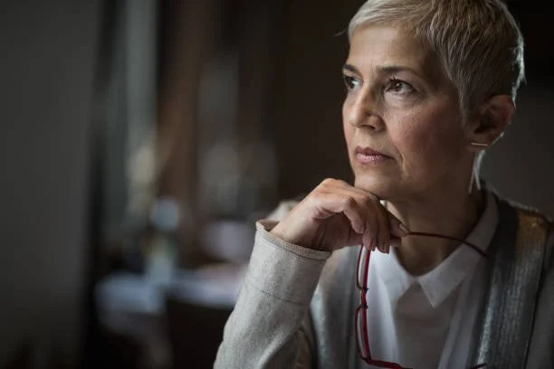 Executive woman leader with short gray hair sits by a window, holding her glasses, looking away in thought.
