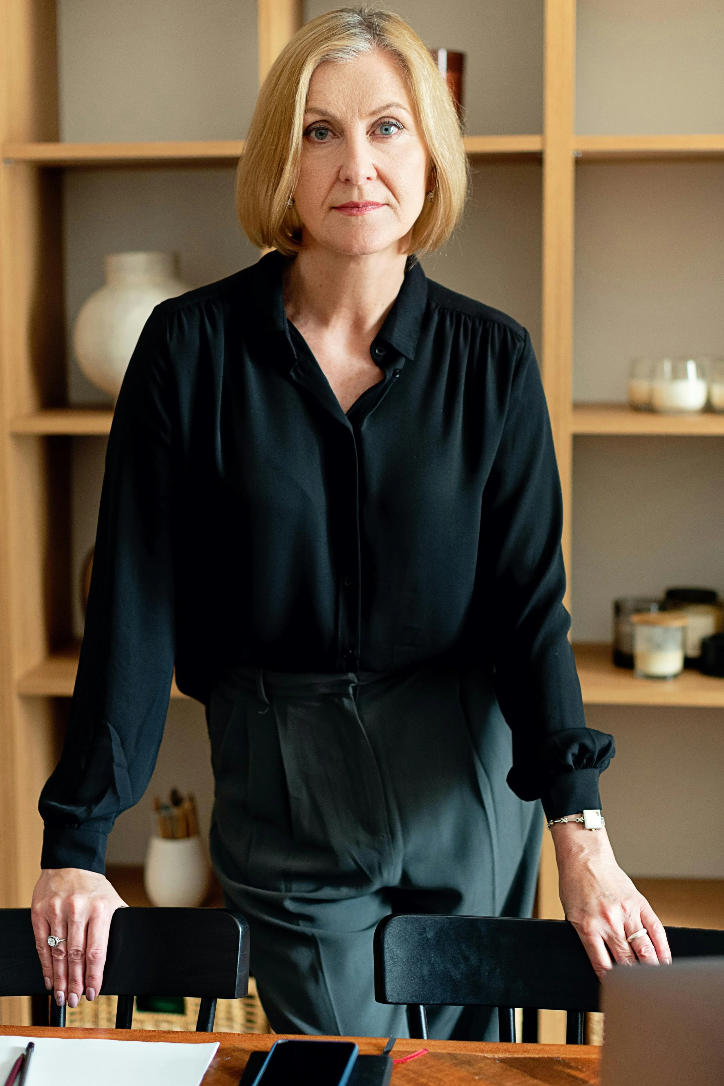 A woman leader with blonde hair in a black blouse stands at a conference table, hands resting on the surface, looking directly at the camera.