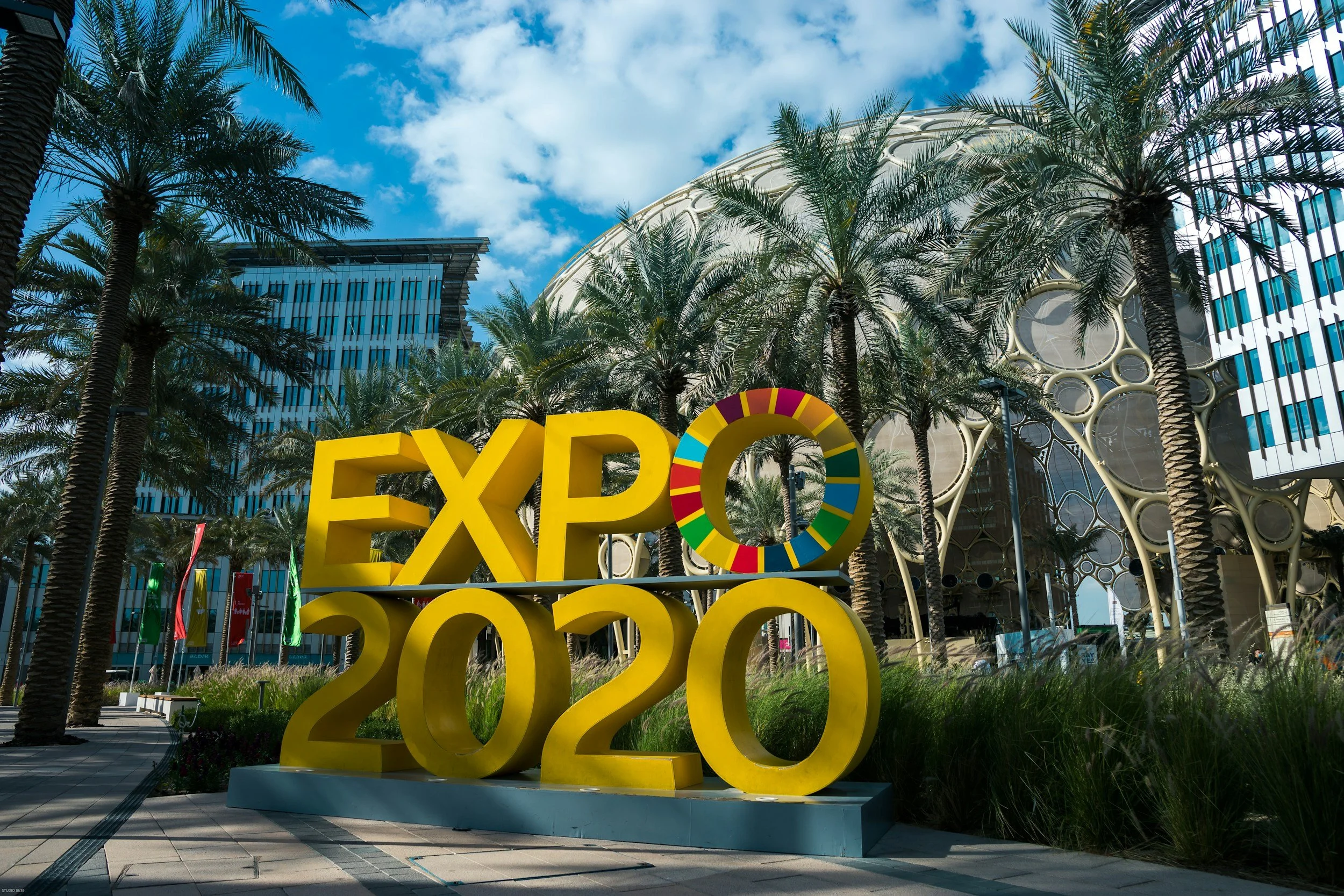 Outdoor scene at the 2022 Expo with large yellow 3D letters spelling 'EXPO' and the year '2022', surrounded by palm trees and modern buildings under a partly cloudy sky.