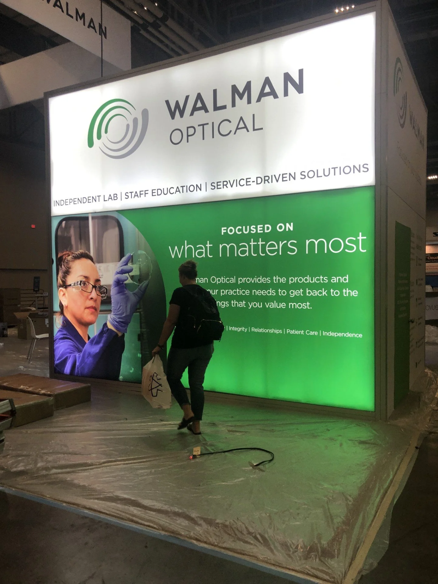 A large illuminated sign at an exhibition booth for Walman Optical displays their logo, tagline, and a woman in protective eyewear inspecting optical lenses. A person with a backpack and shopping bag is walking past.