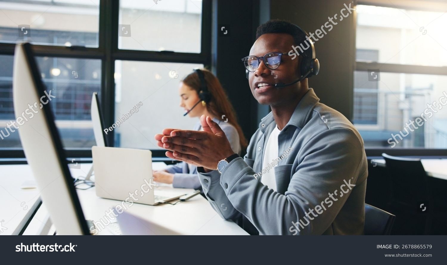 A man wearing glasses and a headset with a microphone sitting at a desk in an office, with a woman working on a laptop in the background.