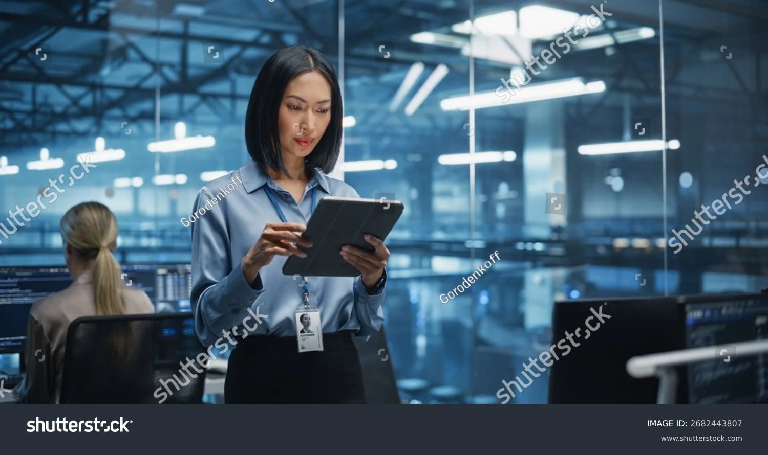 A woman in business attire using a tablet in a modern office with glass walls and computer screens.
