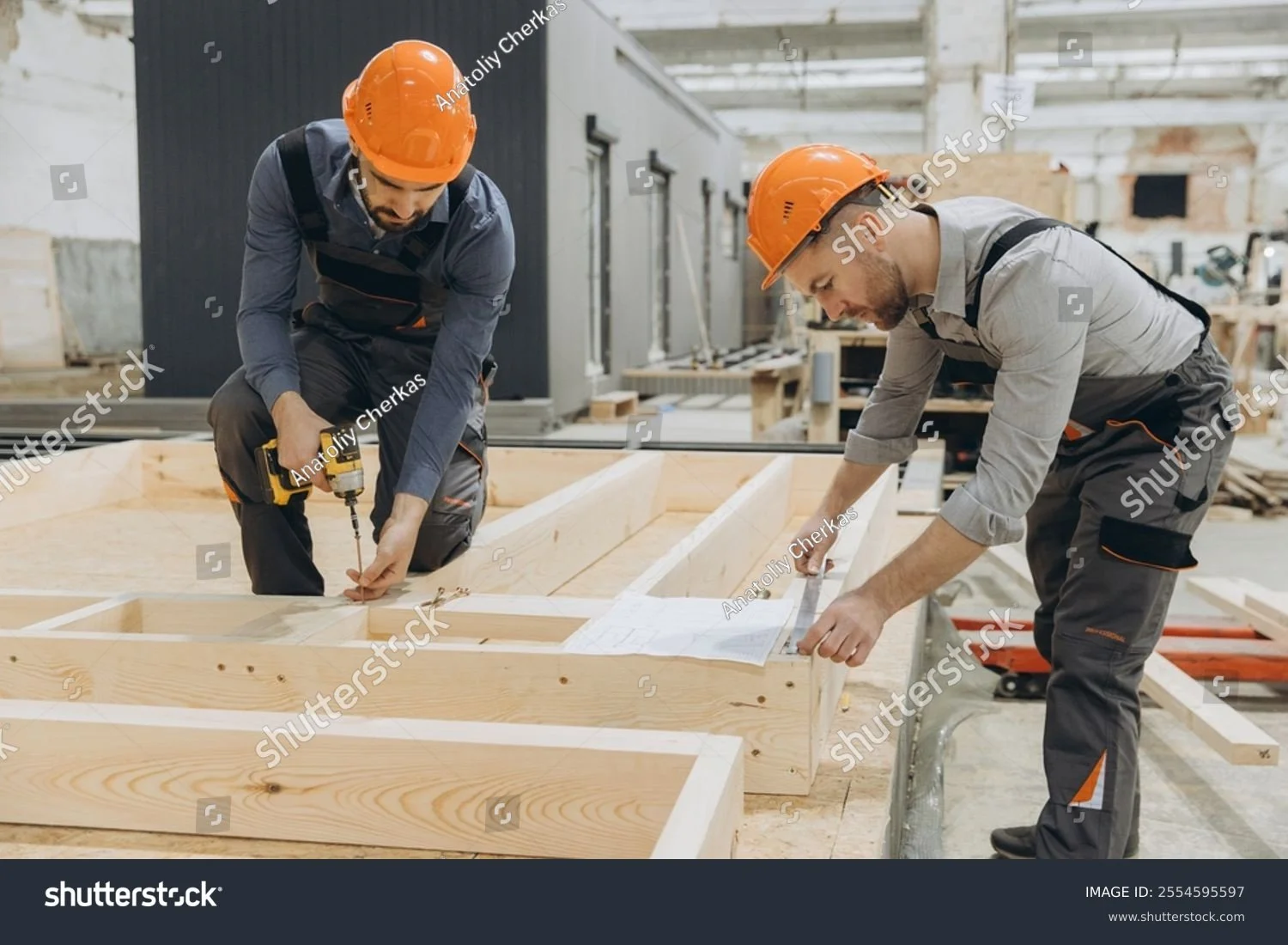 Two construction workers, both wearing orange safety helmets and work clothes, are working on a wooden construction project in a factory or warehouse. One worker is using a cordless drill while the other is measuring with a tape measure and a notepad.