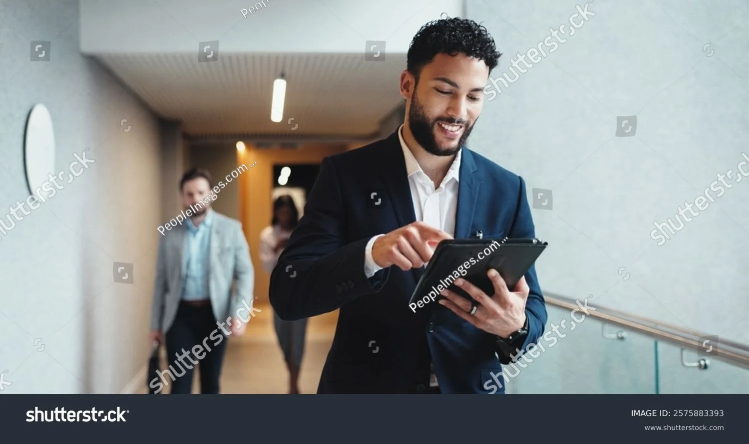 Man in suit using a tablet in an office hallway, with colleagues walking behind him.