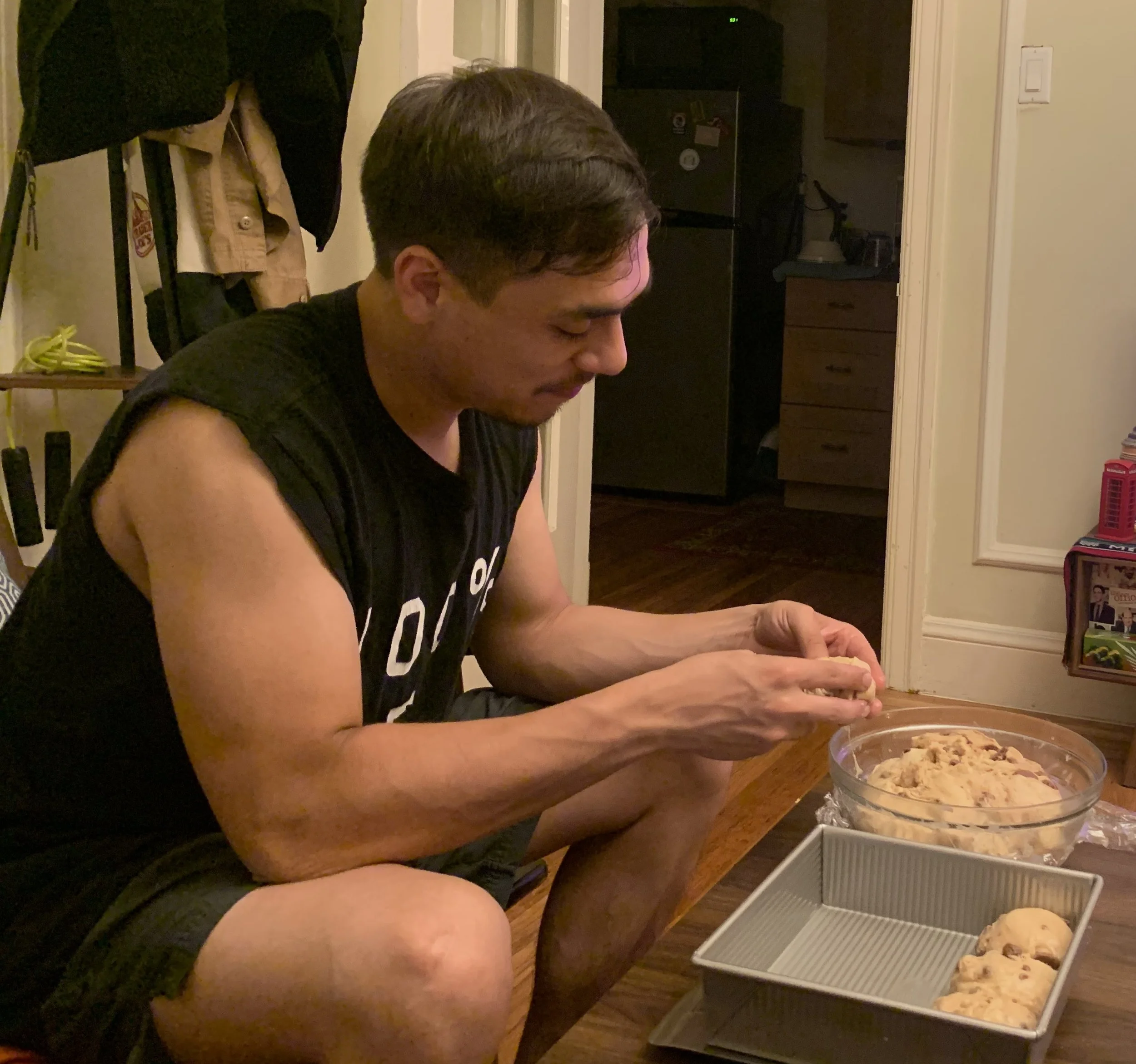 A man in a sleeveless shirt is sitting on the floor, decorating cookies with chocolate chips. There are baking supplies and cookie dough in a glass bowl and a baking pan with cookies nearby.