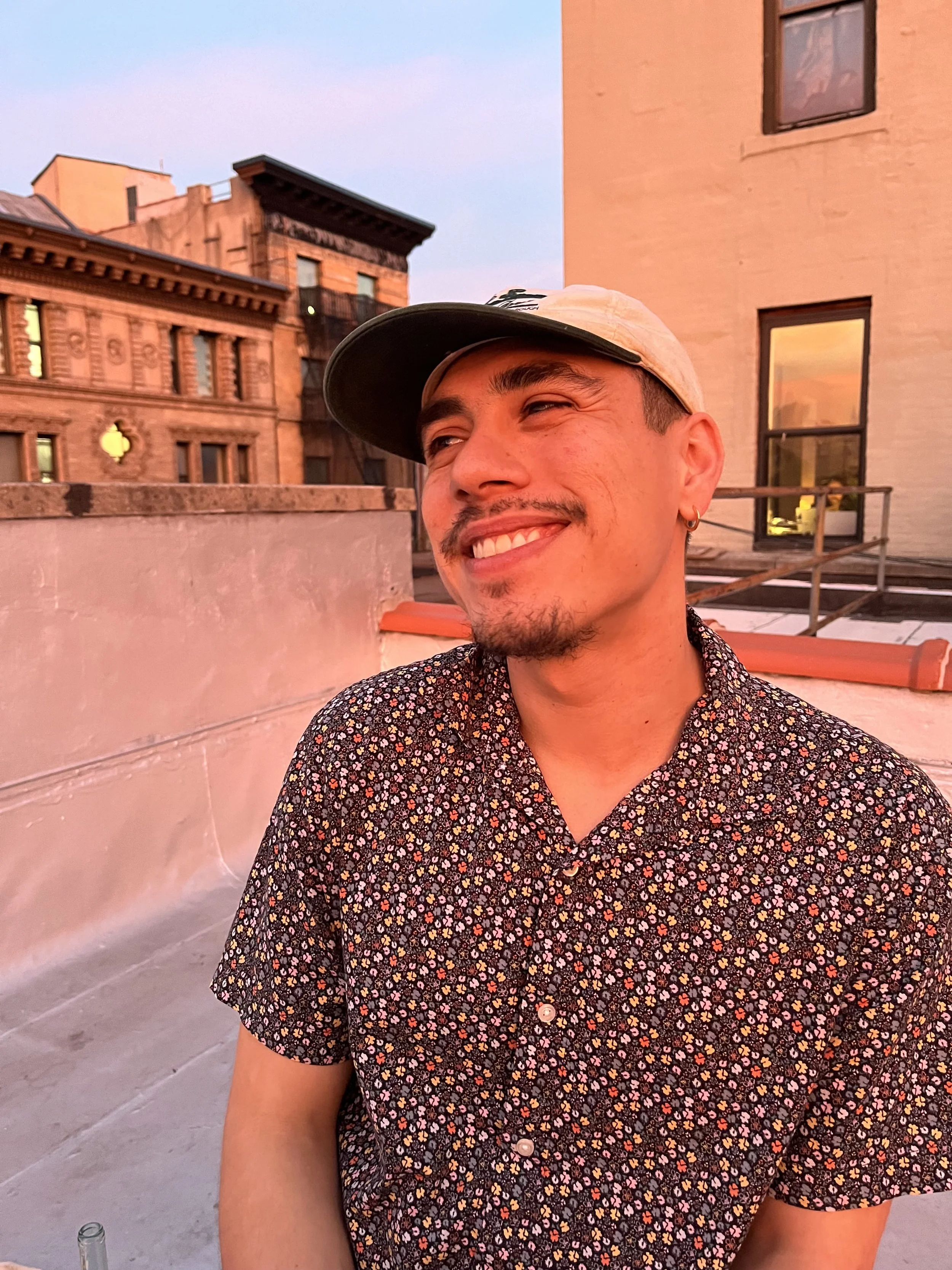 A smiling man in a floral patterned shirt and a beige baseball cap standing on a rooftop during sunset with pink and purple hues in the sky, and buildings in the background.
