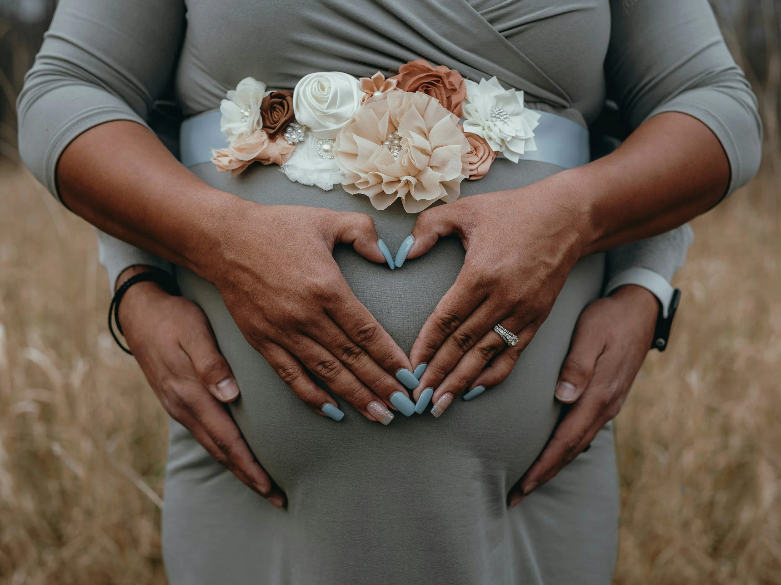 A couple showing their hands and their pregnant belly, with the woman wearing a floral belt and forming a heart shape with their hands.