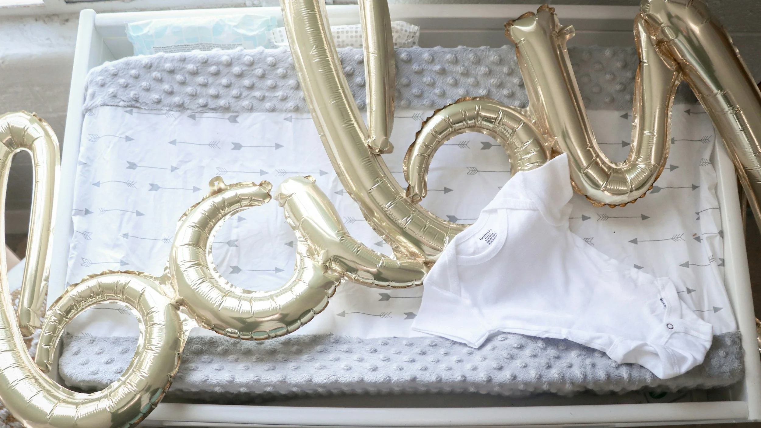 Gold foil balloons spelling out 'baby' placed on a changing table with a gray blanket, a white baby onesie, and a patterned paper underneath, likely for a baby shower celebration.