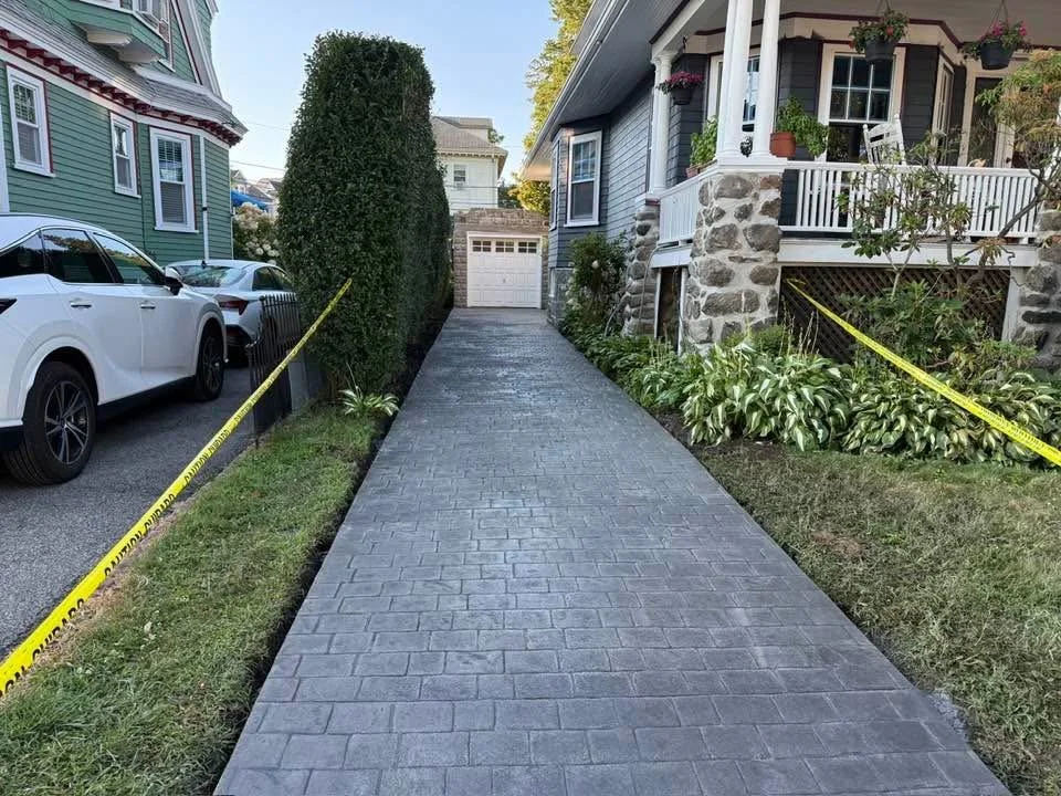 Concrete driveway leading to a garage, flanked by a manicured lawn and plants on both sides, with parked cars and a house on the right.