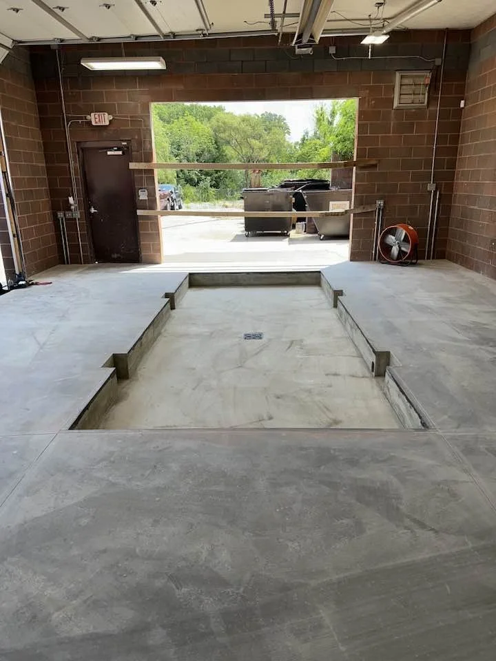 The image shows an indoor space under construction, with an open rectangular area in the floor that appears to be for a vehicle lift or pit, surrounded by concrete edges. The room has brick walls, a closed door on the left, and an open passageway at the back leading outside to a green, tree-filled area. Construction materials and tools are visible, including a ventilation fan on the right.