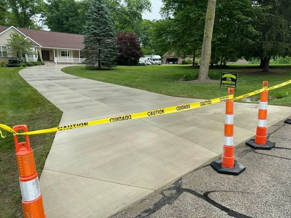 Concrete driveway section cordoned off with yellow caution tape and orange traffic cones in front of a house, with trees, lawn, and a sign advertising concrete services.