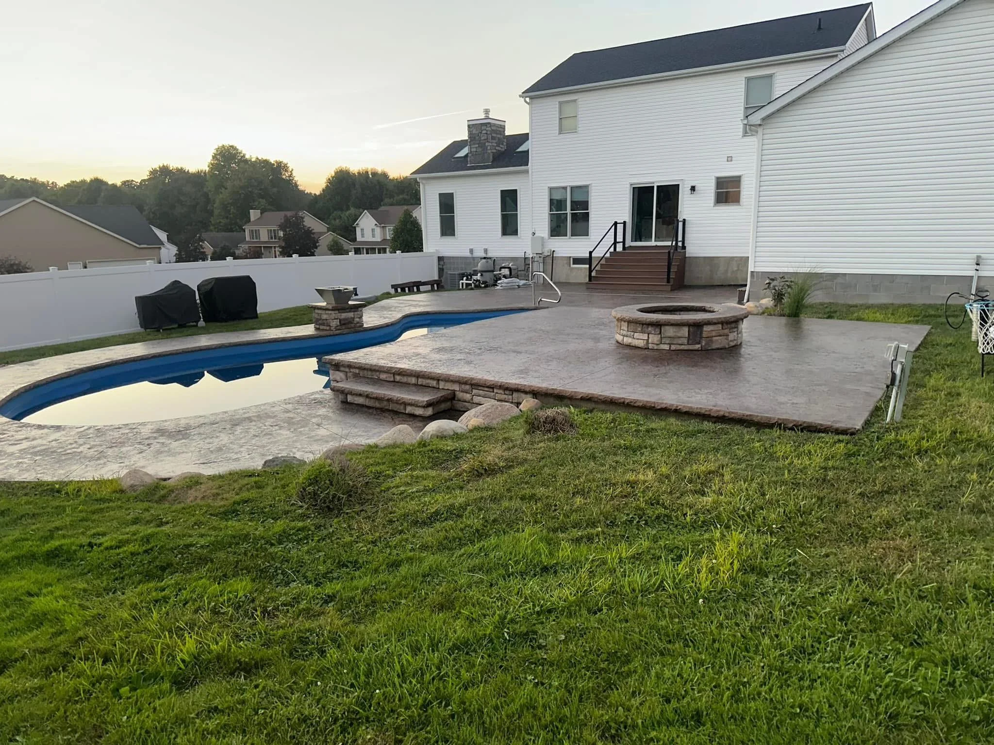 Backyard with a kidney-shaped swimming pool, a raised fire pit, and a concrete patio attached to a white house with sliding glass door and stairs. The yard is fenced, and there are outdoor furniture and grill covers.