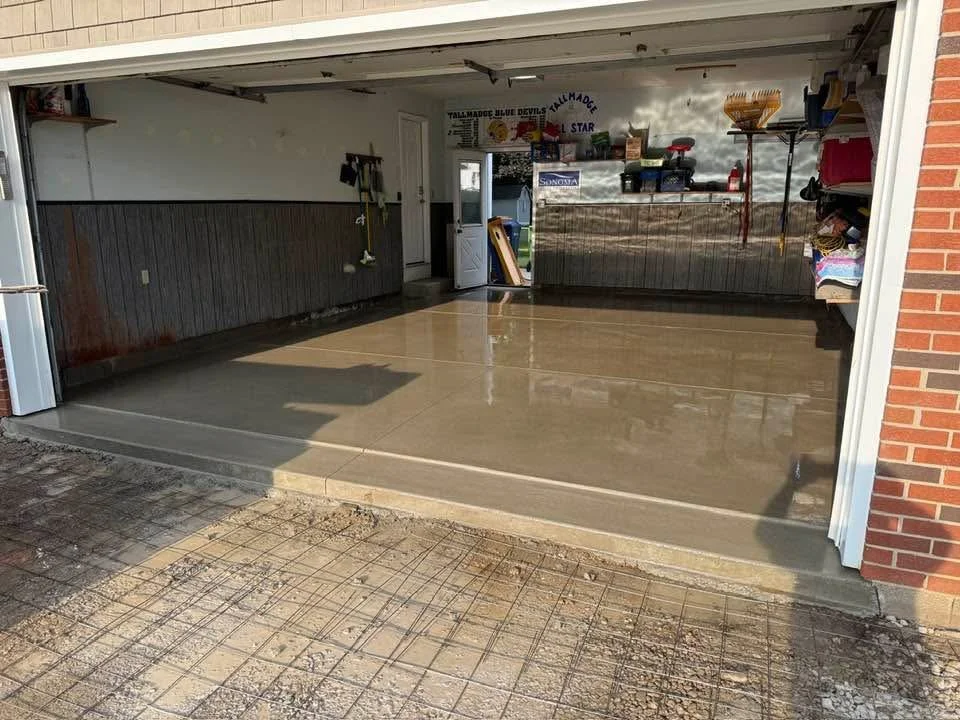 A newly poured and smooth concrete garage floor inside an open garage with tools and yard supplies on shelves and on wall