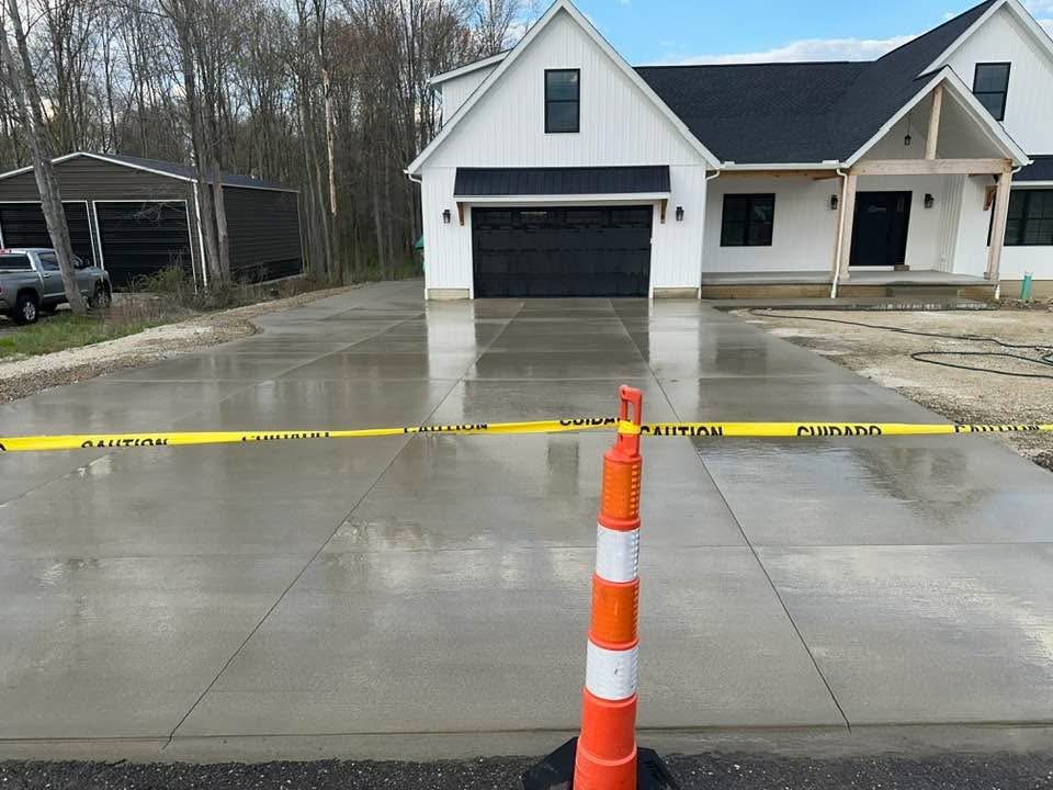 Newly paved and wet concrete driveway in front of a white house with black garage door, caution tape, and an orange traffic cone.