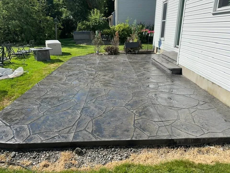 Recently poured concrete patio with a decorative stamped pattern next to a white house and green grass, with garden plants and outdoor furniture nearby.