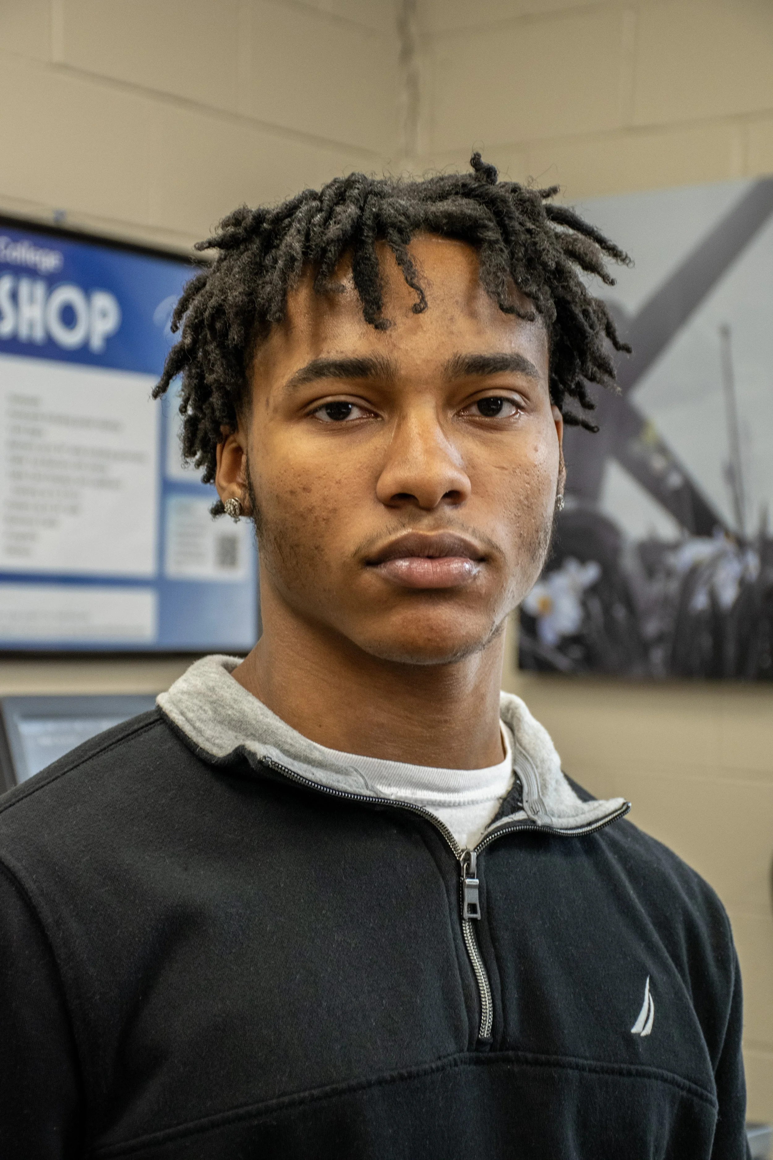A young man with dark, textured hair and earrings, wearing a black zip-up jacket over a gray shirt, standing indoors, with a blurred background and a sign on the wall.