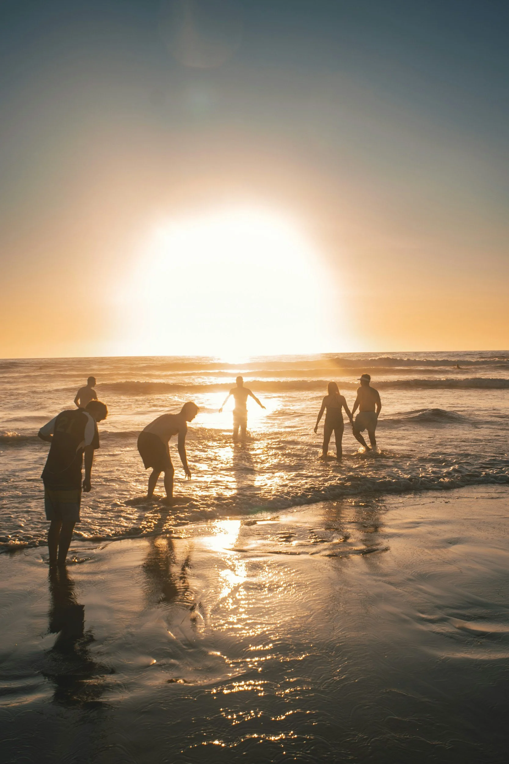 People enjoying a sunset at the beach, with some wading in the water and holding hands, silhouettes reflected in the ocean. Samara, Costa Rica