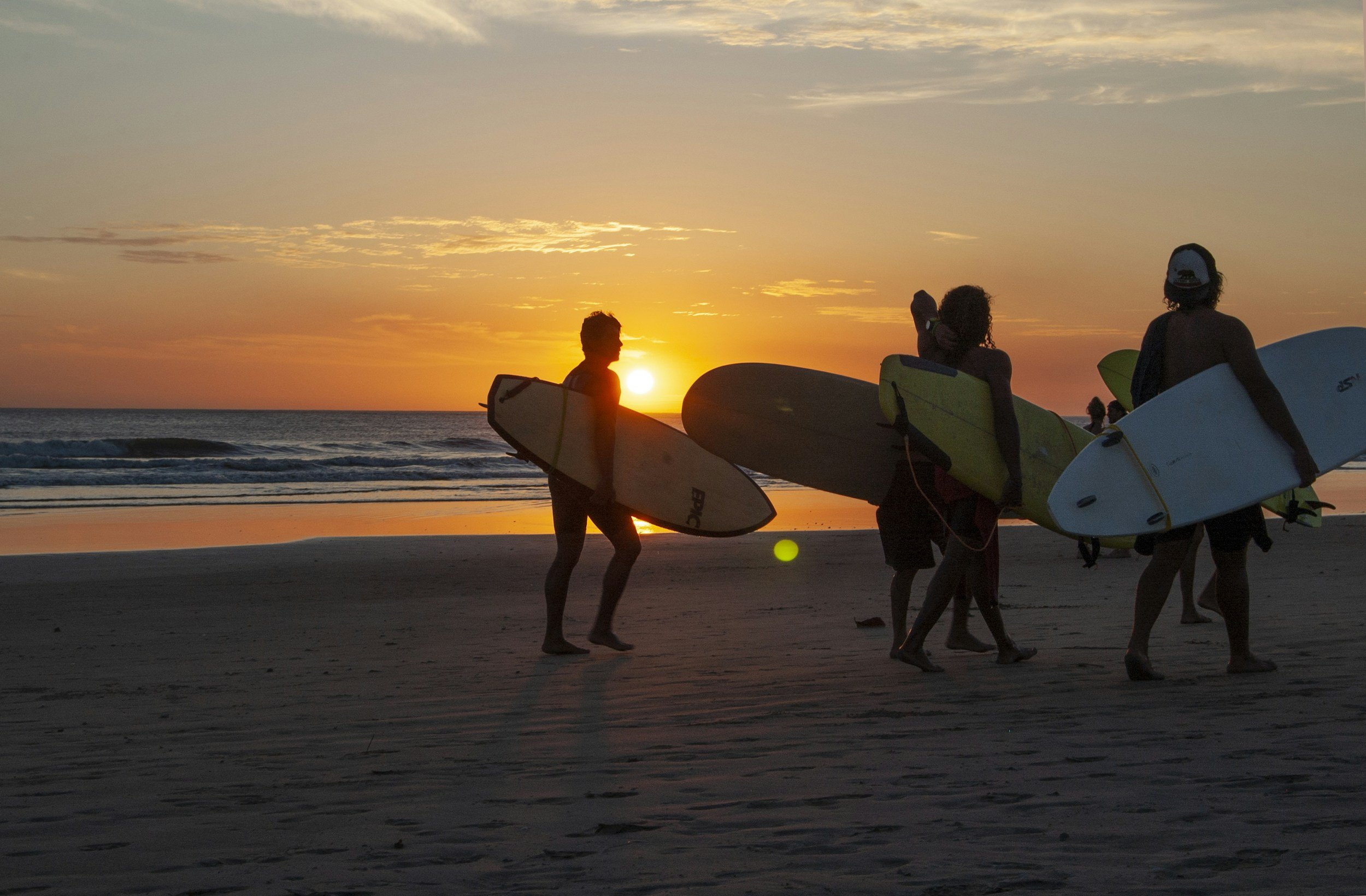 Silhouettes of people carrying surfboards walking on a beach at sunset, with the sun near the horizon and a cloudy sky. Samara, Costa Rica