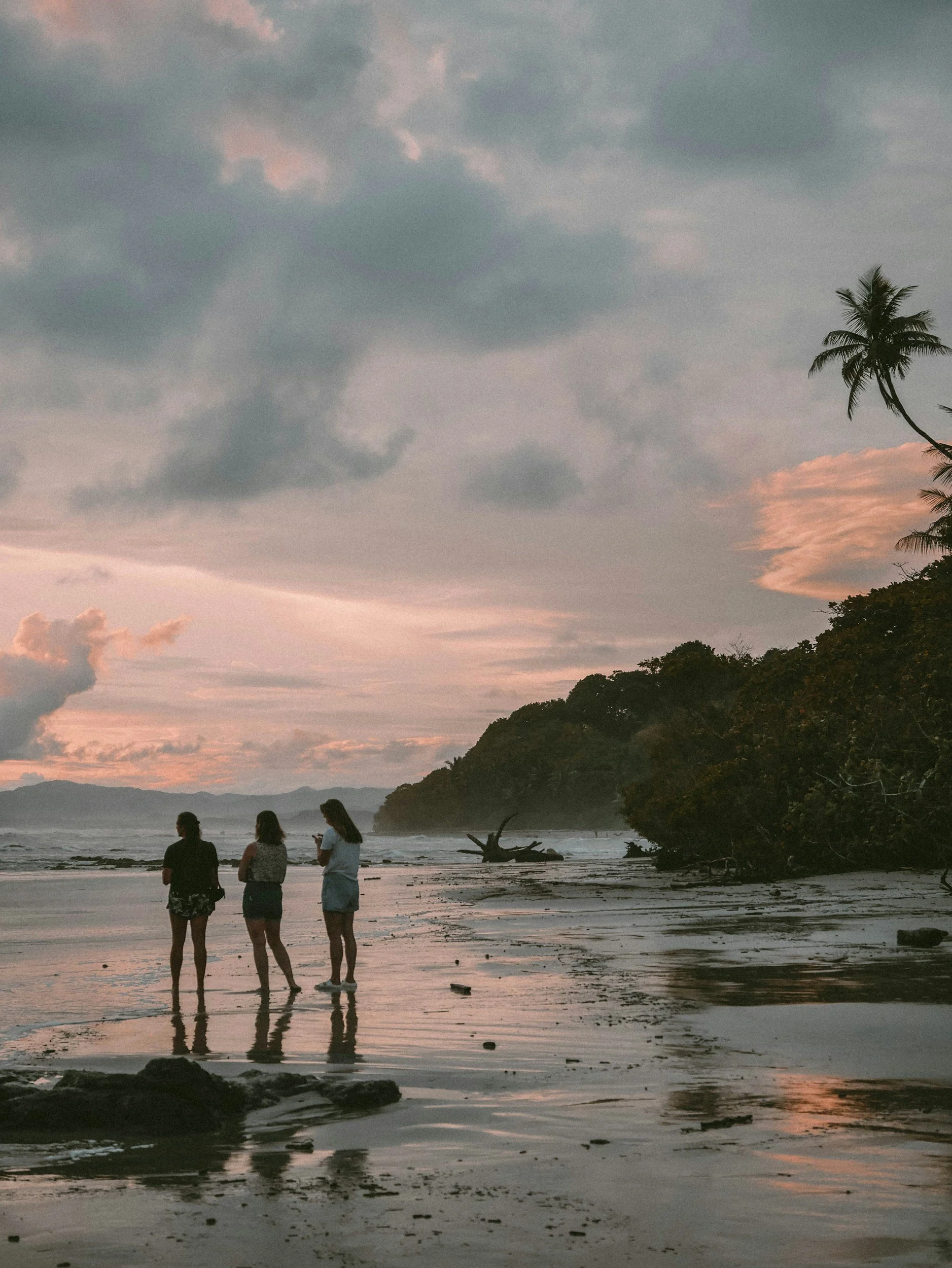 Three people standing on the beach at sunset, with clouds in the sky and a large tree on the right side of the image. Samara, Costa Rica
