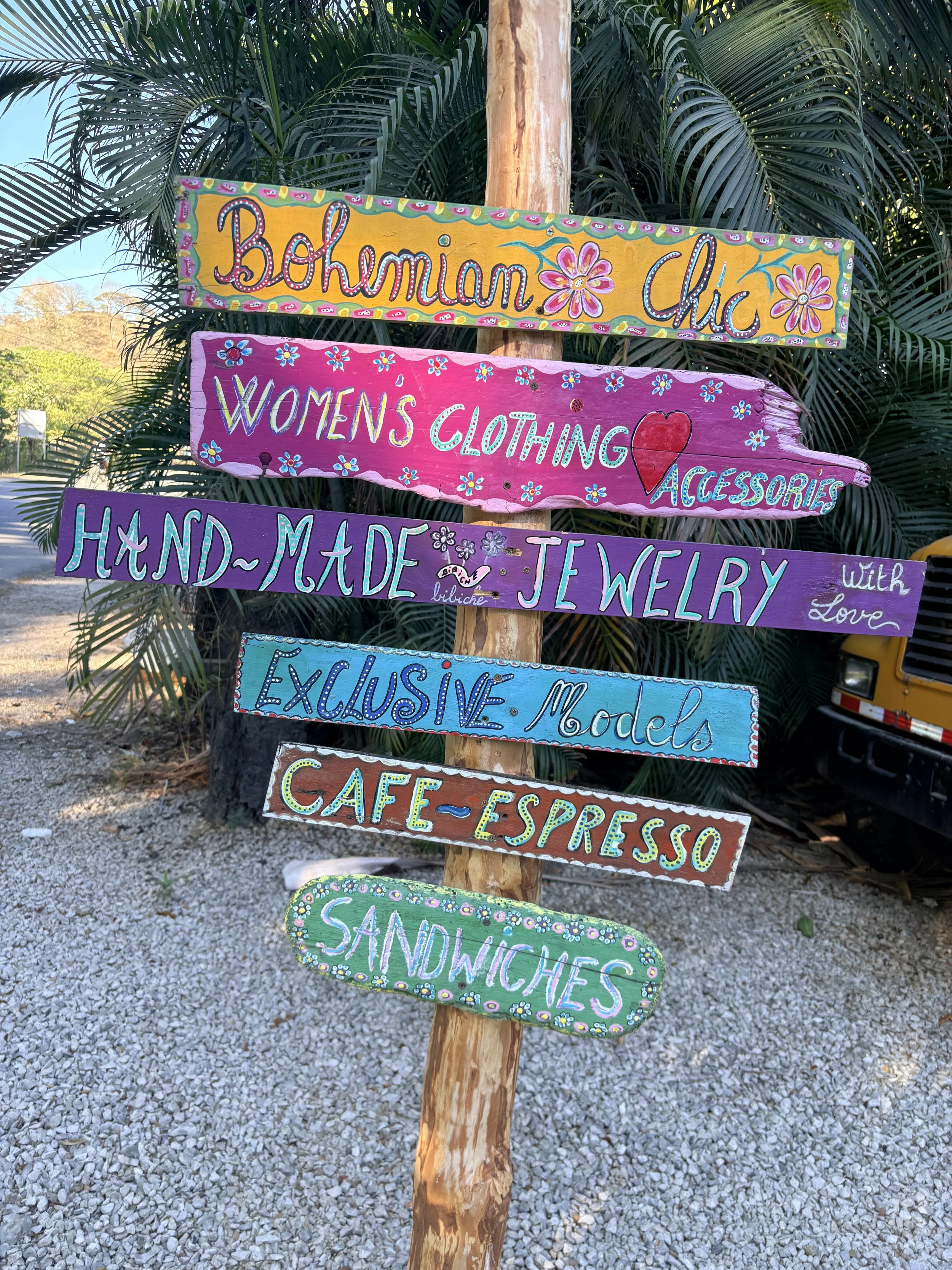 Colorful hand-painted wooden signs on a tree advertising shops selling women's clothing, accessories, jewelry, and sandwiches, with tropical palm leaves in the background. Samara, Costa Rica