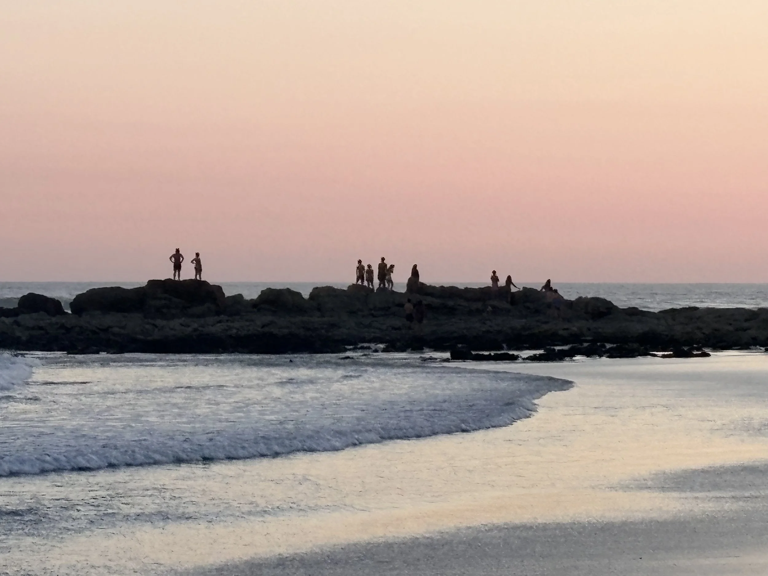 People standing and sitting on rocks along a beach at sunset, with the ocean waves in the foreground and a pastel pink sky in the background. Samara, Costa Rica