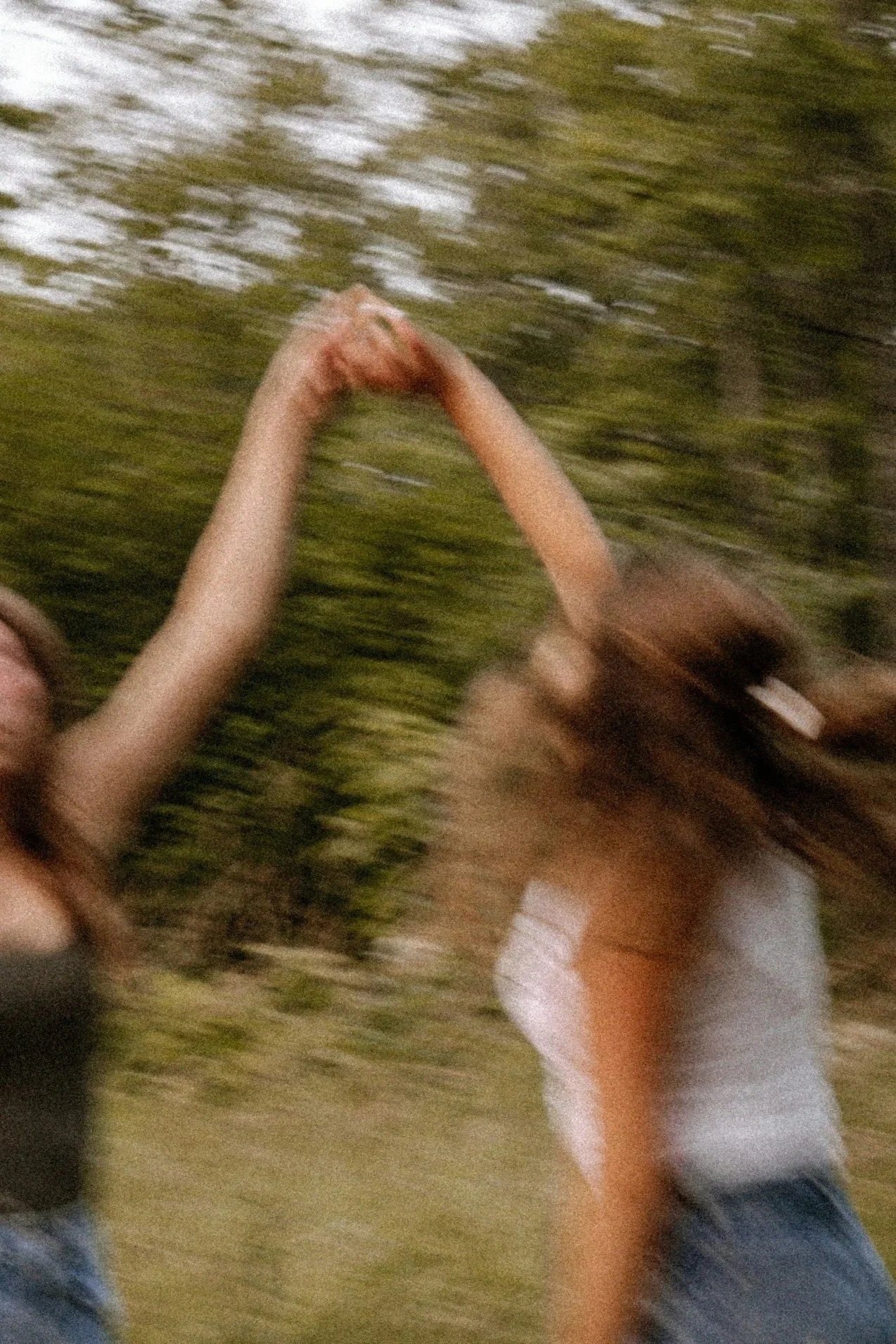 Two women holding hands while spinning or dancing outdoors on a grassy area with trees in the background.