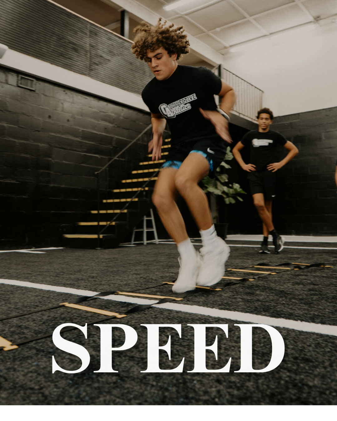 Young man with curly hair jump training on speed agility ladder, with another person in the background in an indoor gym.