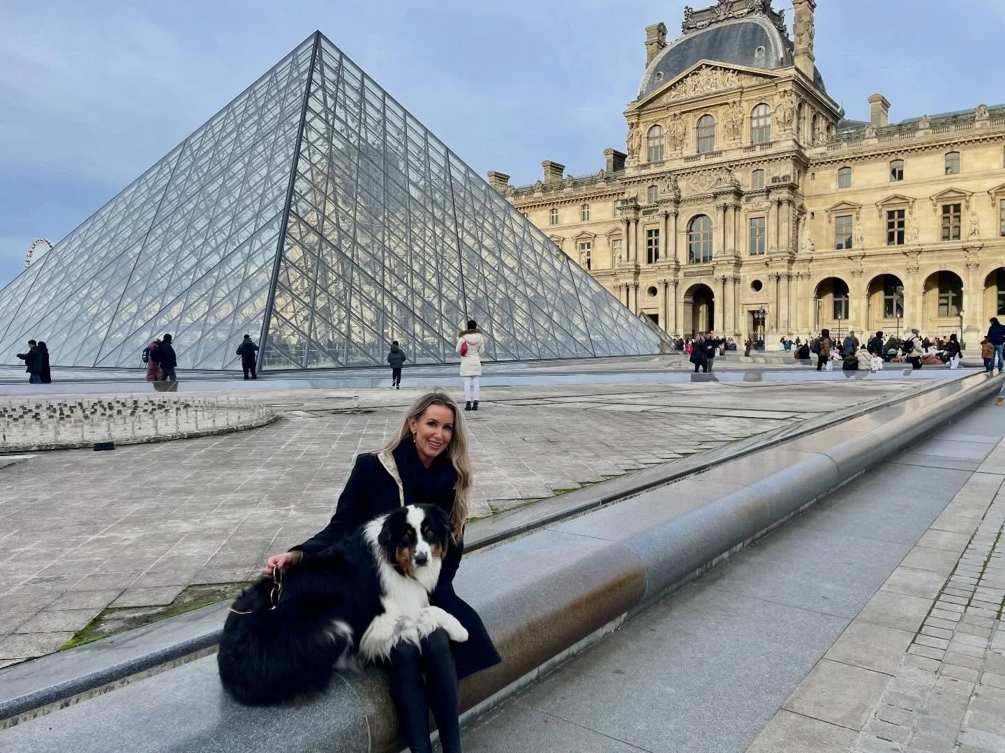 A woman sitting on a stone bench with her Australian Shepherd dog in front of the Louvre Pyramid and historic building in Paris, France, on a clear day.