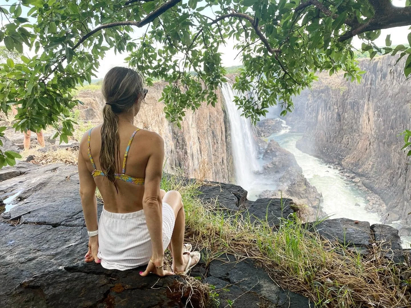 Woman sitting on rocks near a waterfall in a lush green setting over looking Victoria Falls.
