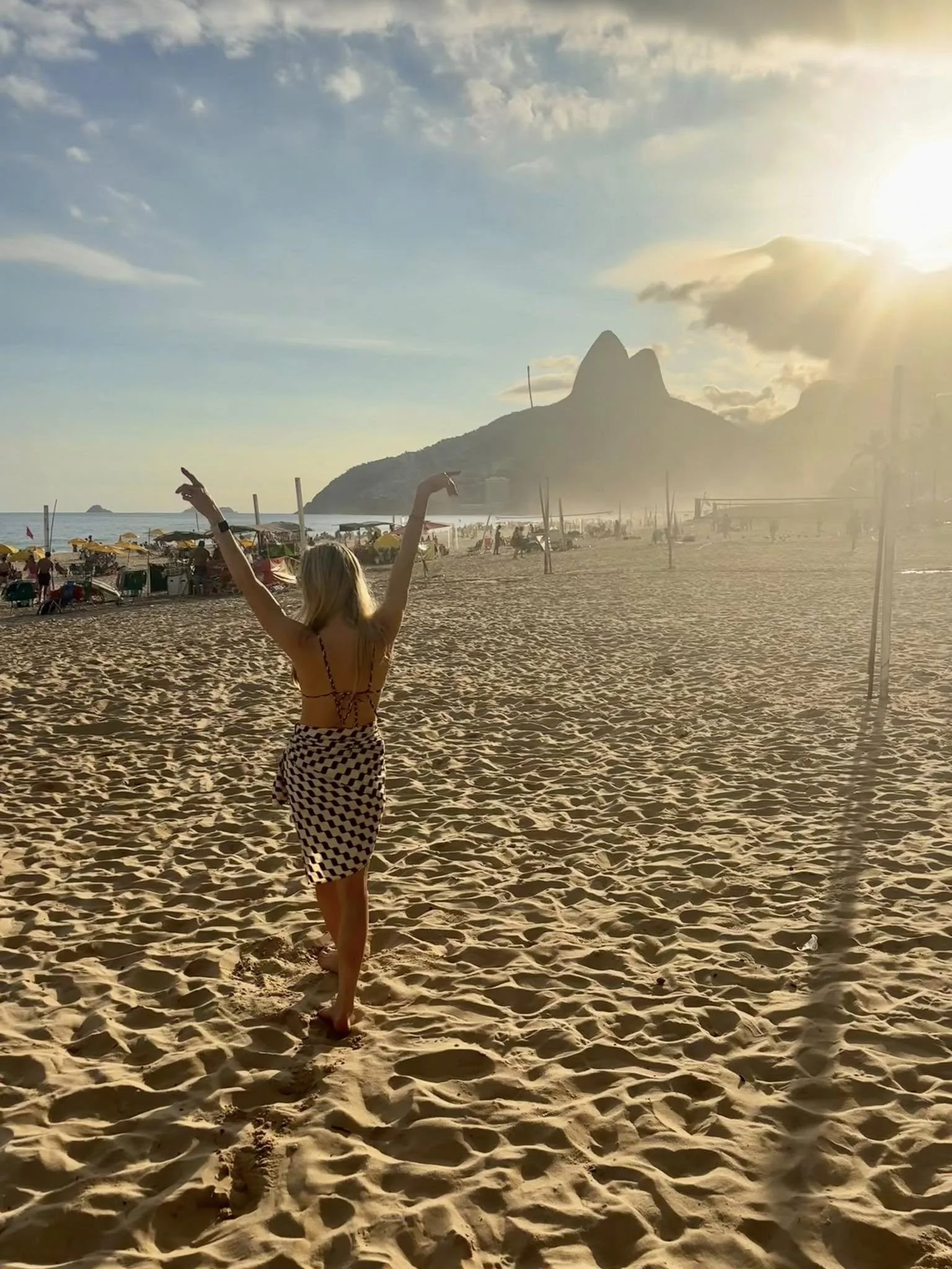 A woman walking on the beach during sunset, with Rio de Janeiro's Sugarloaf Mountain in the background.