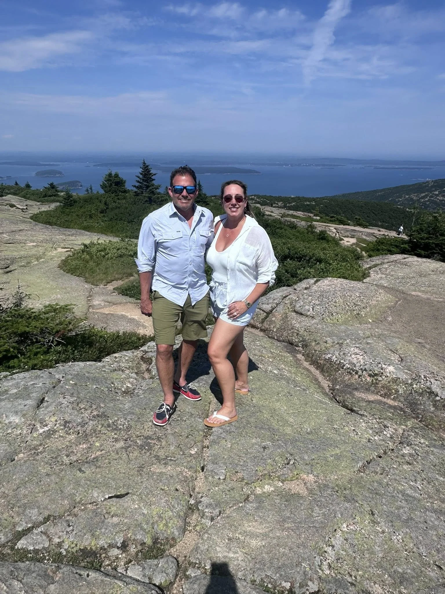 A smiling man and woman standing on a rocky mountain summit with a scenic view of a lake, islands, and a partly cloudy sky behind them.