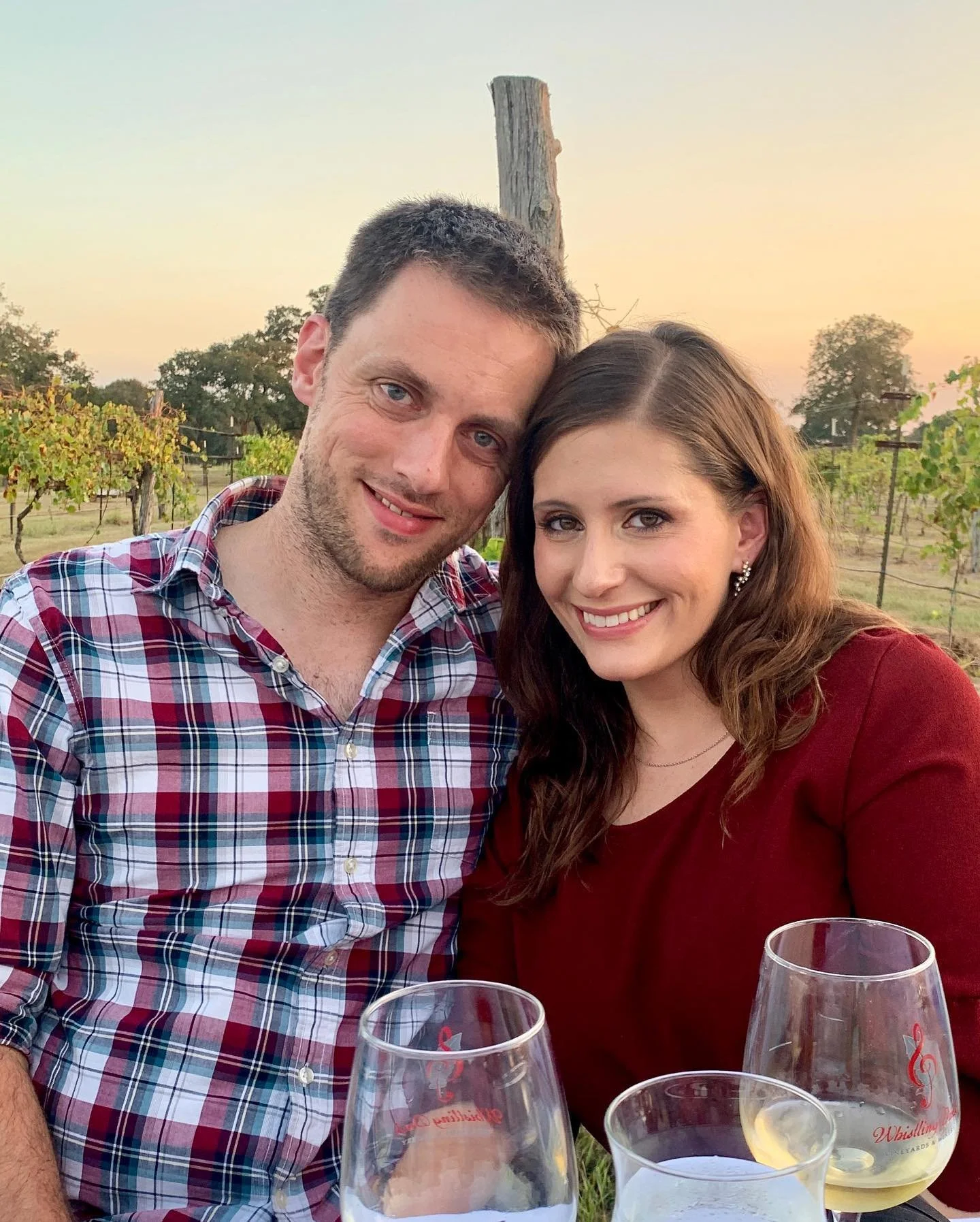 A smiling couple sitting outdoors at sunset with wine glasses in front of them, a vineyard in the background.
