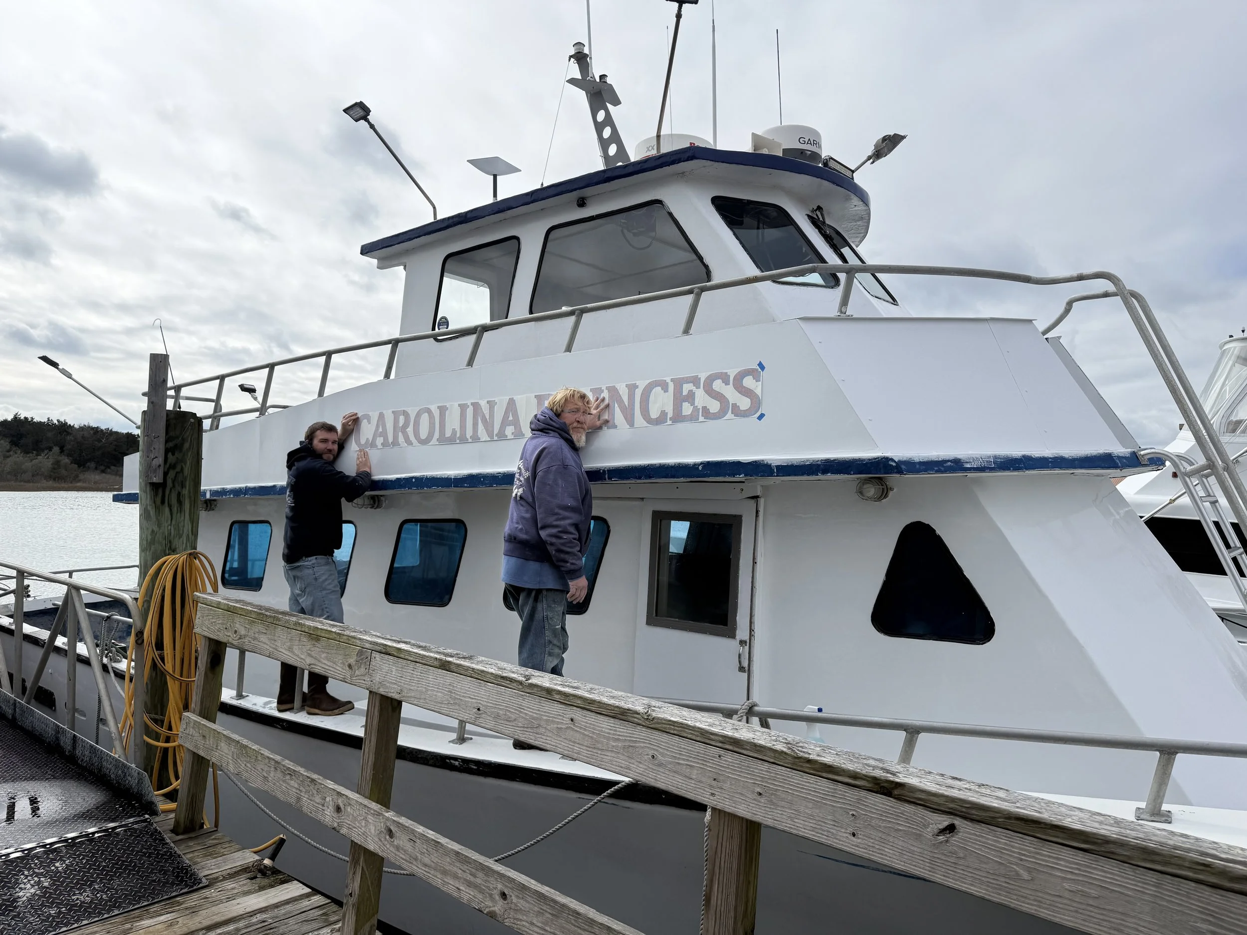 A white boat named Carolina Princess docked at a pier, with two men standing nearby. One man has blond hair and a beard, wearing a gray hoodie, and the other man is wearing a black jacket, with the background showing water and cloudy sky.