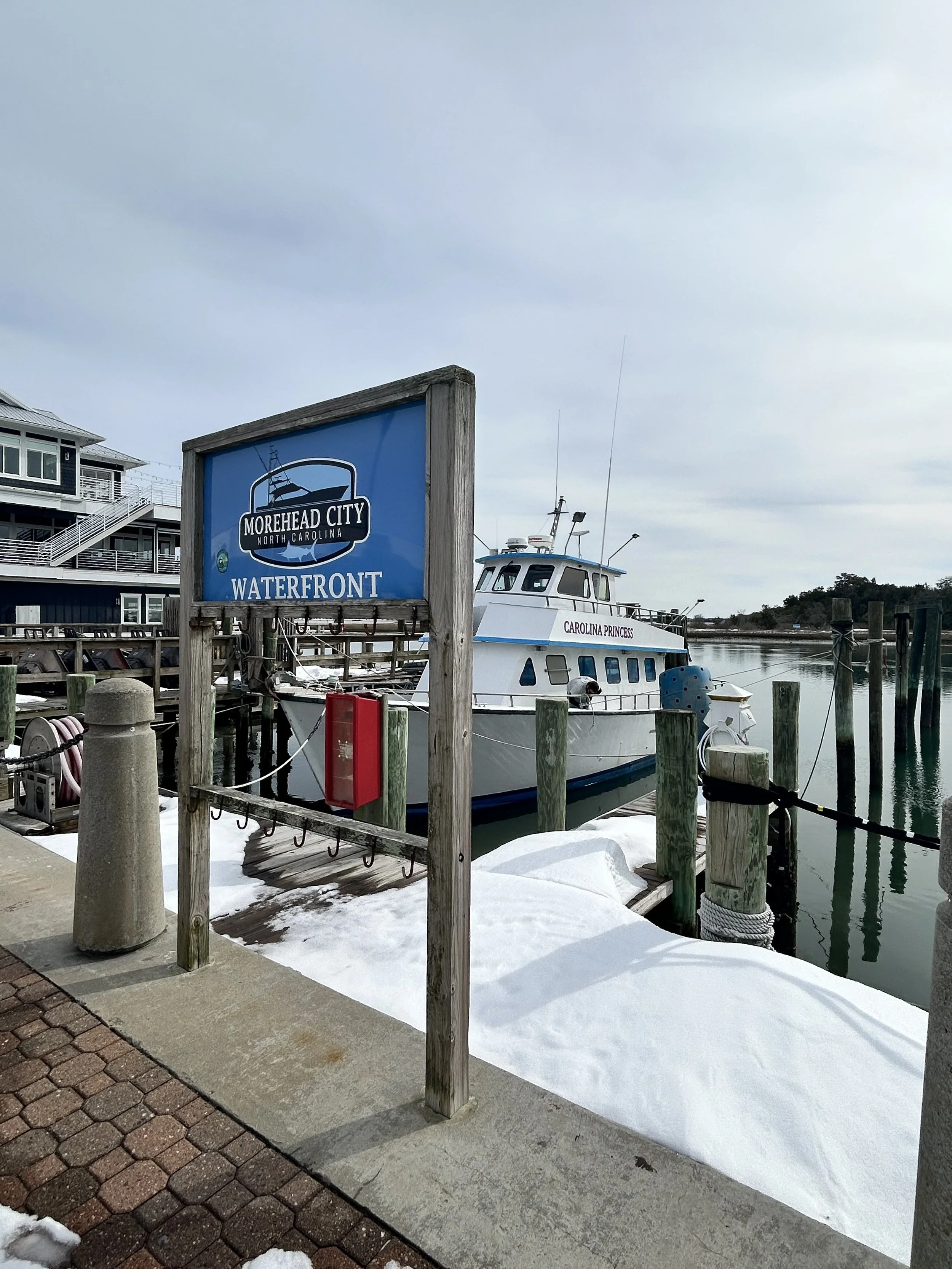 A marina with boats docked, a sign reading 'Morehead City North Carolina Waterfront,' snow on the ground, and a cloudy sky.