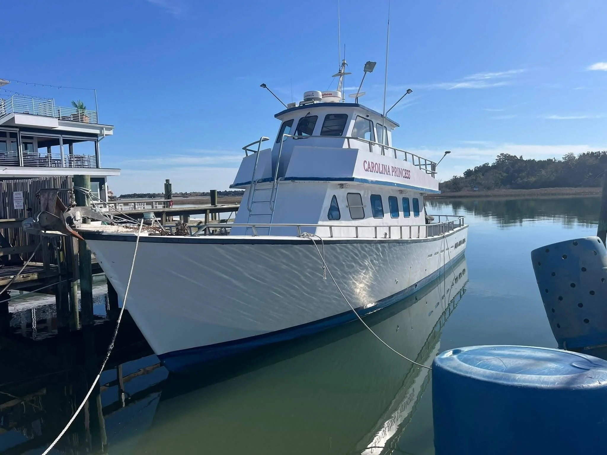 White boat named Carolina Princess docked near a wooden pier on calm water, with a clear blue sky and trees in the background.
