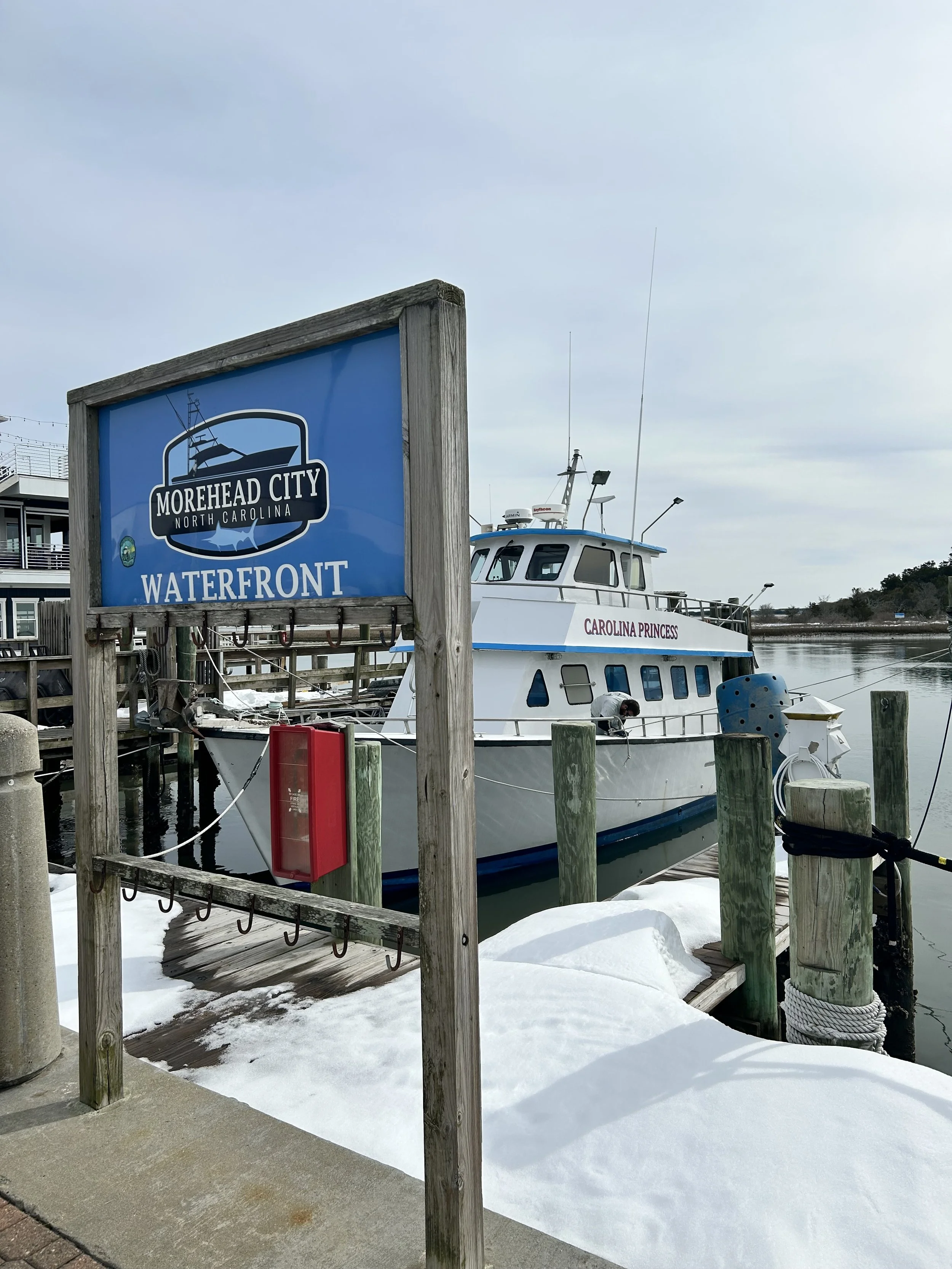 A marina with a boat named Carolina Princess docked, surrounded by snow on the ground, with a blue sign indicating Morehead City Waterfront in North Carolina.