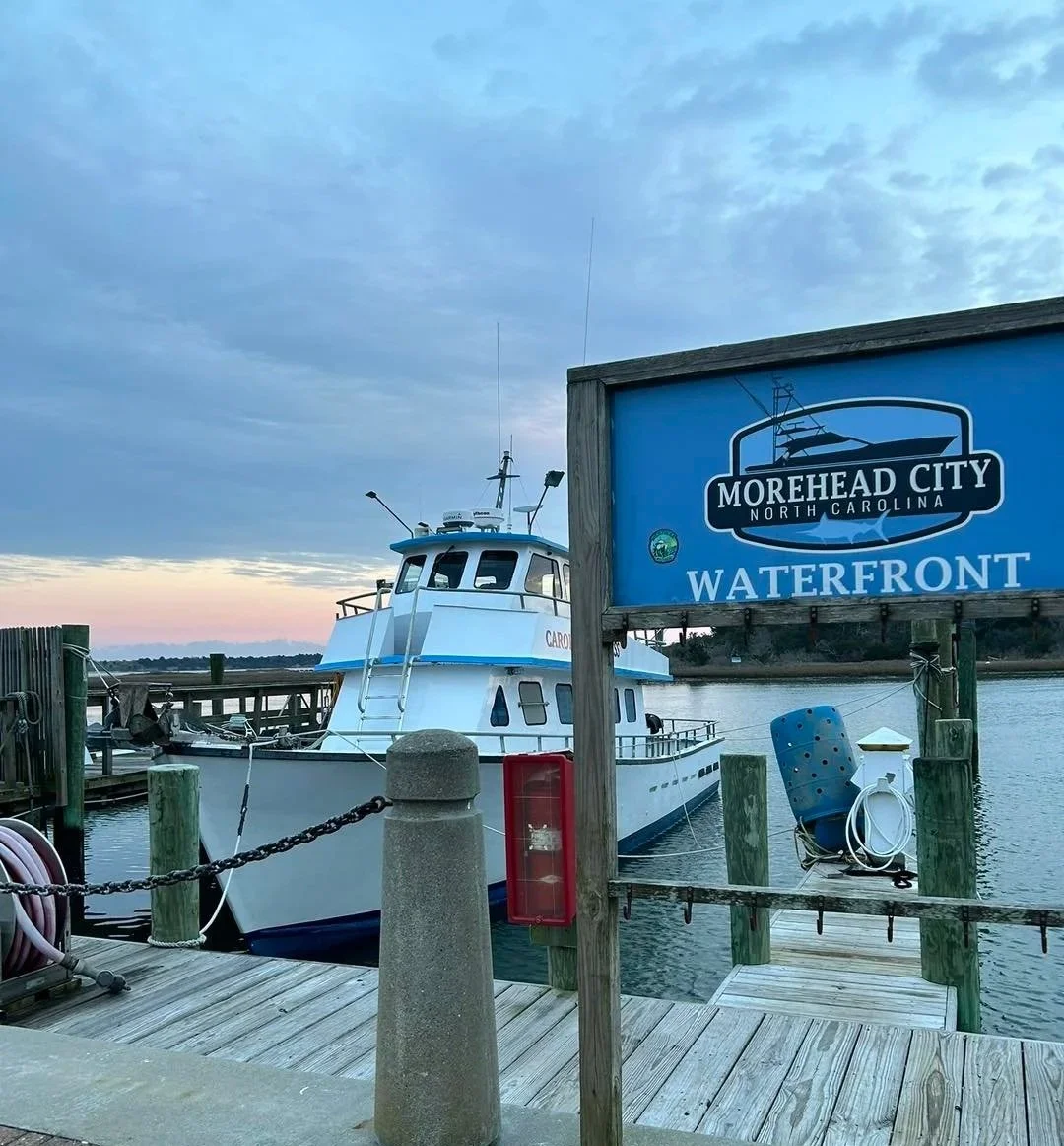 A boat docked at a waterfront in Morehead City, North Carolina, with a sign indicating the location and a cloudy sky in the background.