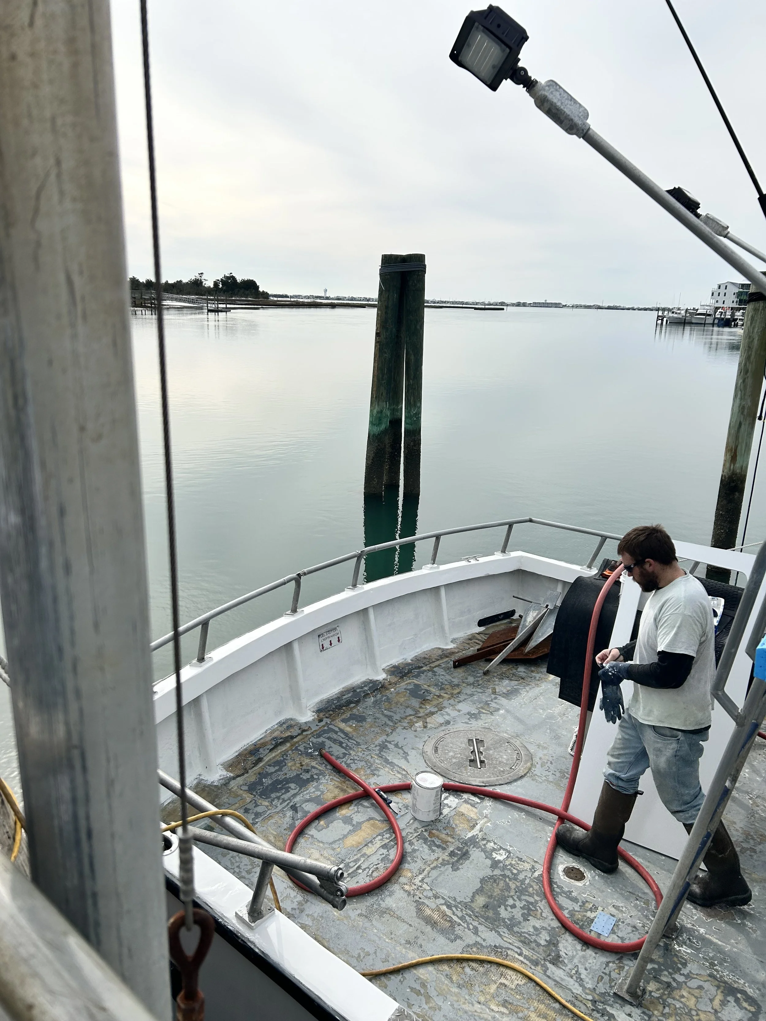 A man standing on a boat deck wearing rubber boots, a long sleeve shirt, and gloves, working with hoses. The river and docked boats are visible in the background under a cloudy sky.