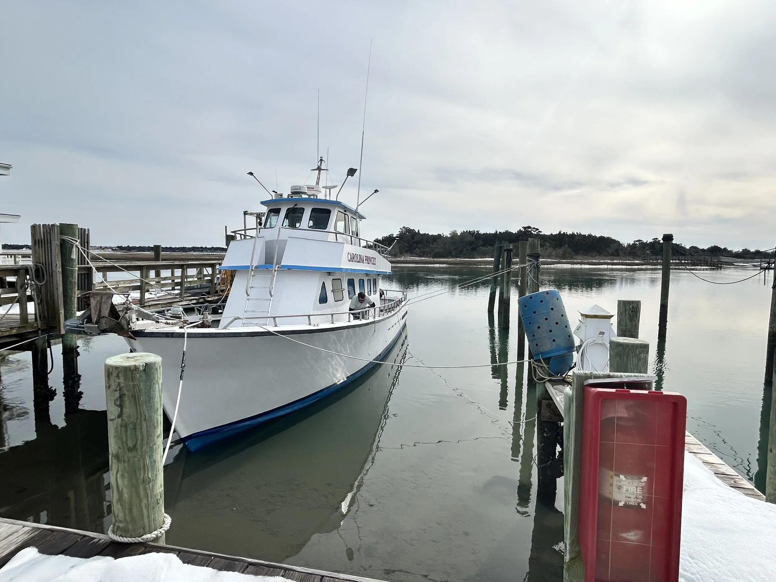 A white boat named Carolina Princess docked at a marina with calm water and cloudy sky, surrounded by wooden posts and ropes.