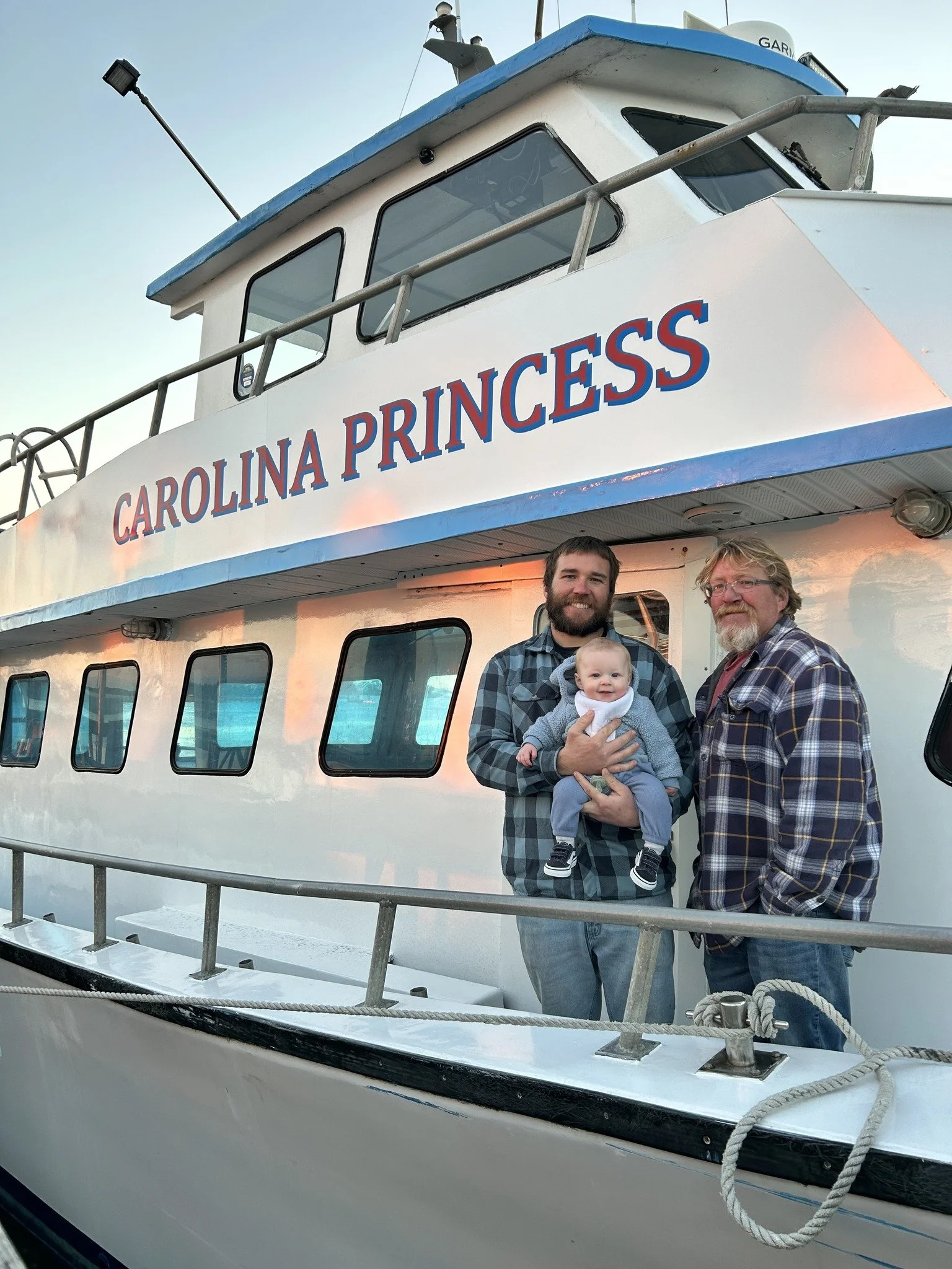 Two men and a baby standing on a boat named Carolina Princess, posing for a photo.