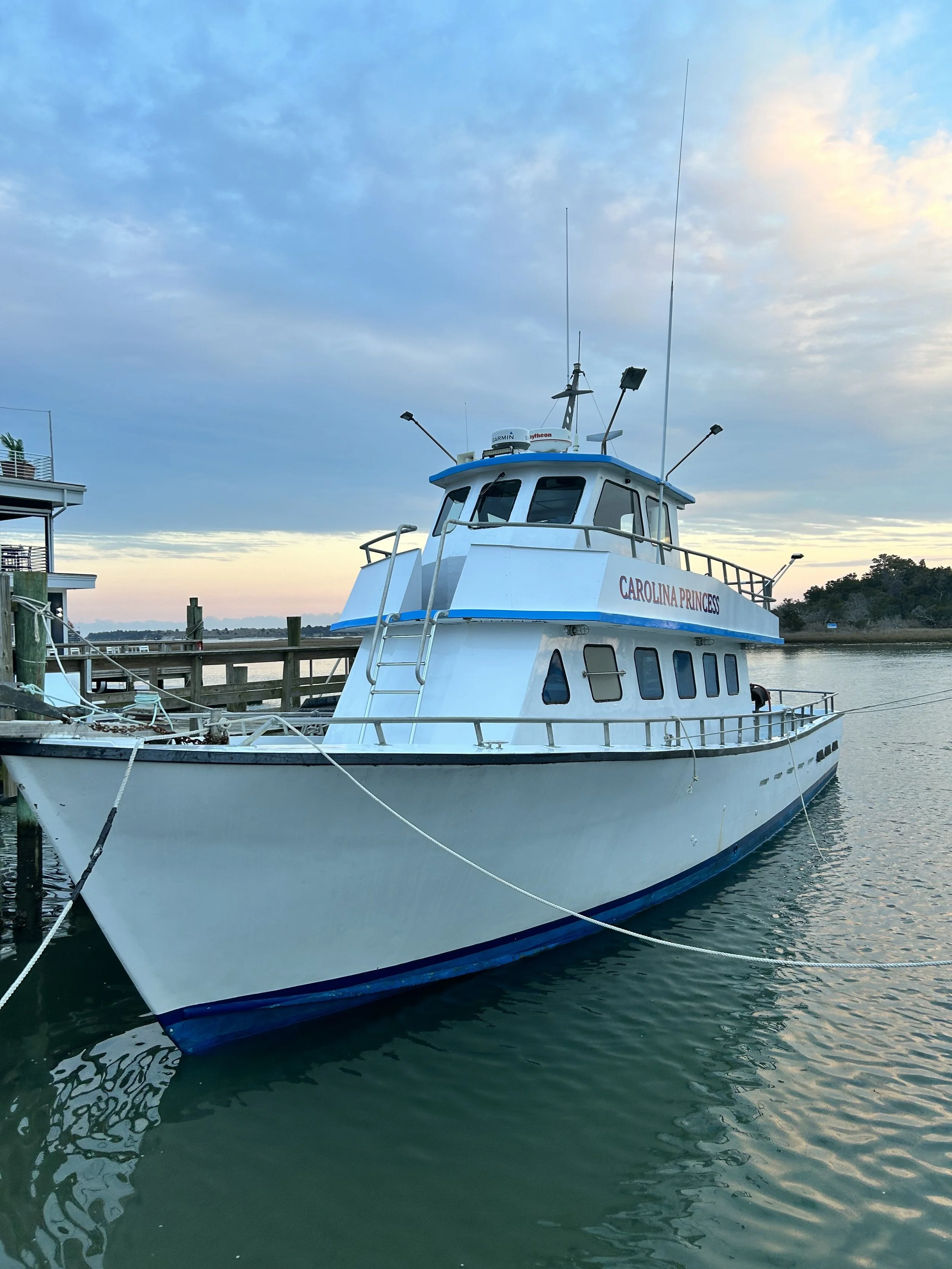 A white boat named 'Carolina Princess' docked at a pier on a calm body of water during sunset, with blue sky and scattered clouds in the background.