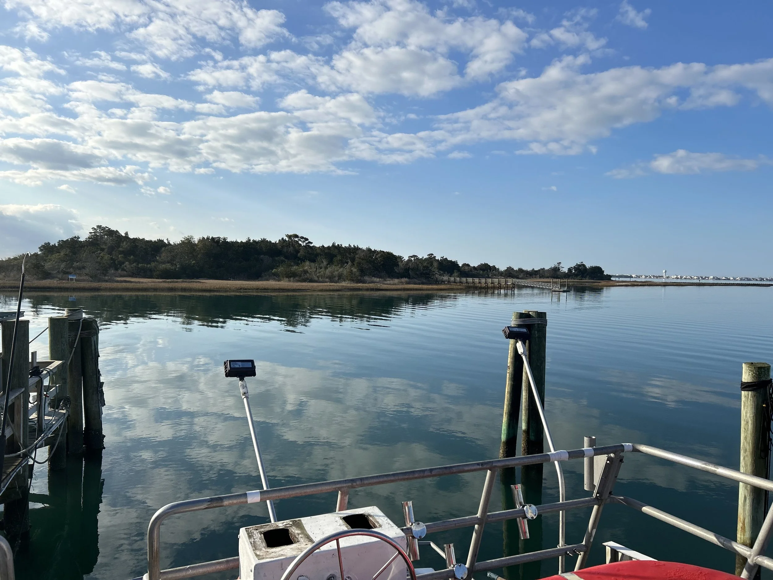 View of a calm body of water with trees and a shoreline in the background under a partly cloudy sky, taken from a dock or boat with wooden posts and equipment visible in the foreground.