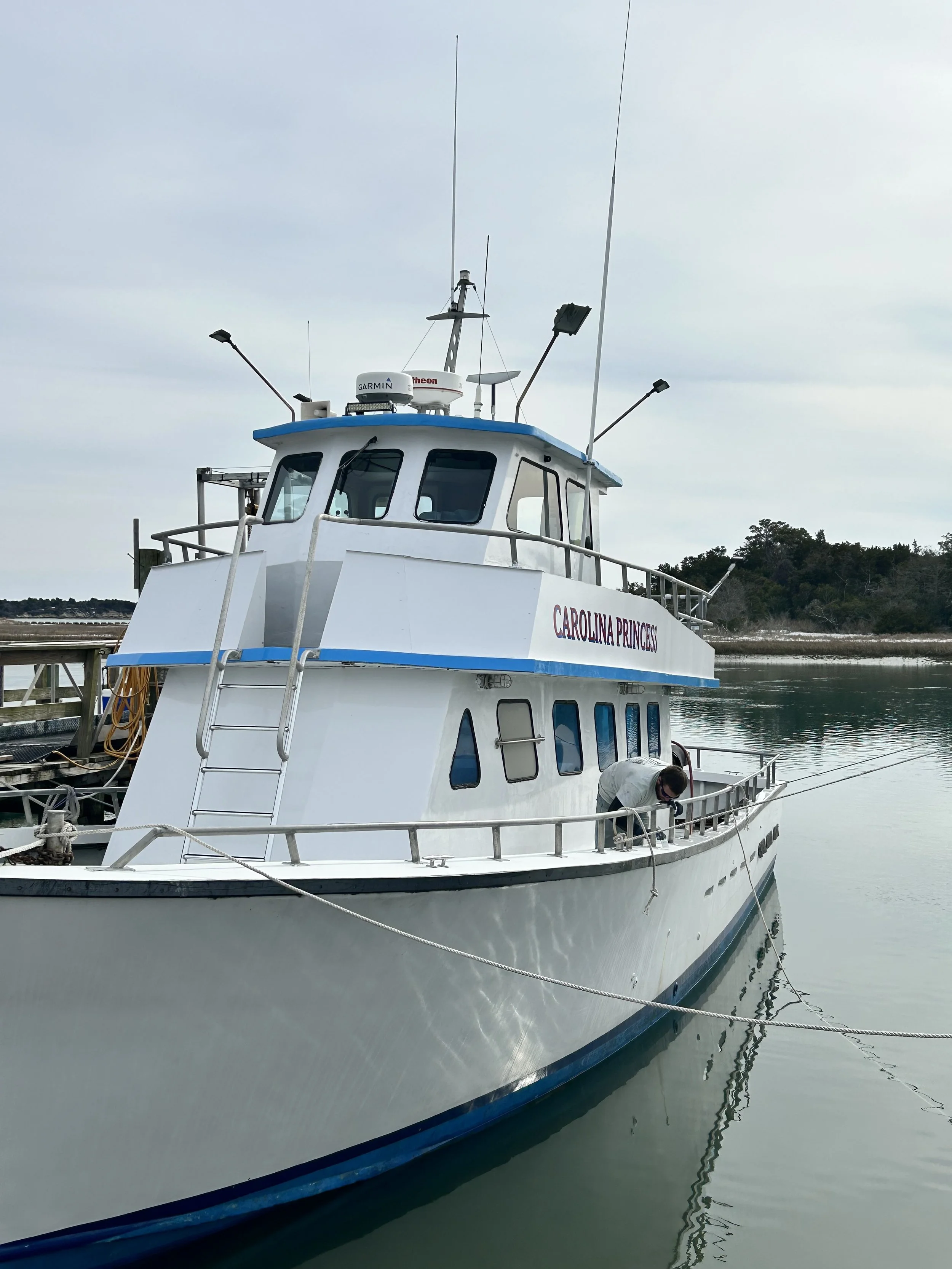A white boat docked at a marina with calm water, trees in the background, and equipment on the boat including radar, antennas, and lights.