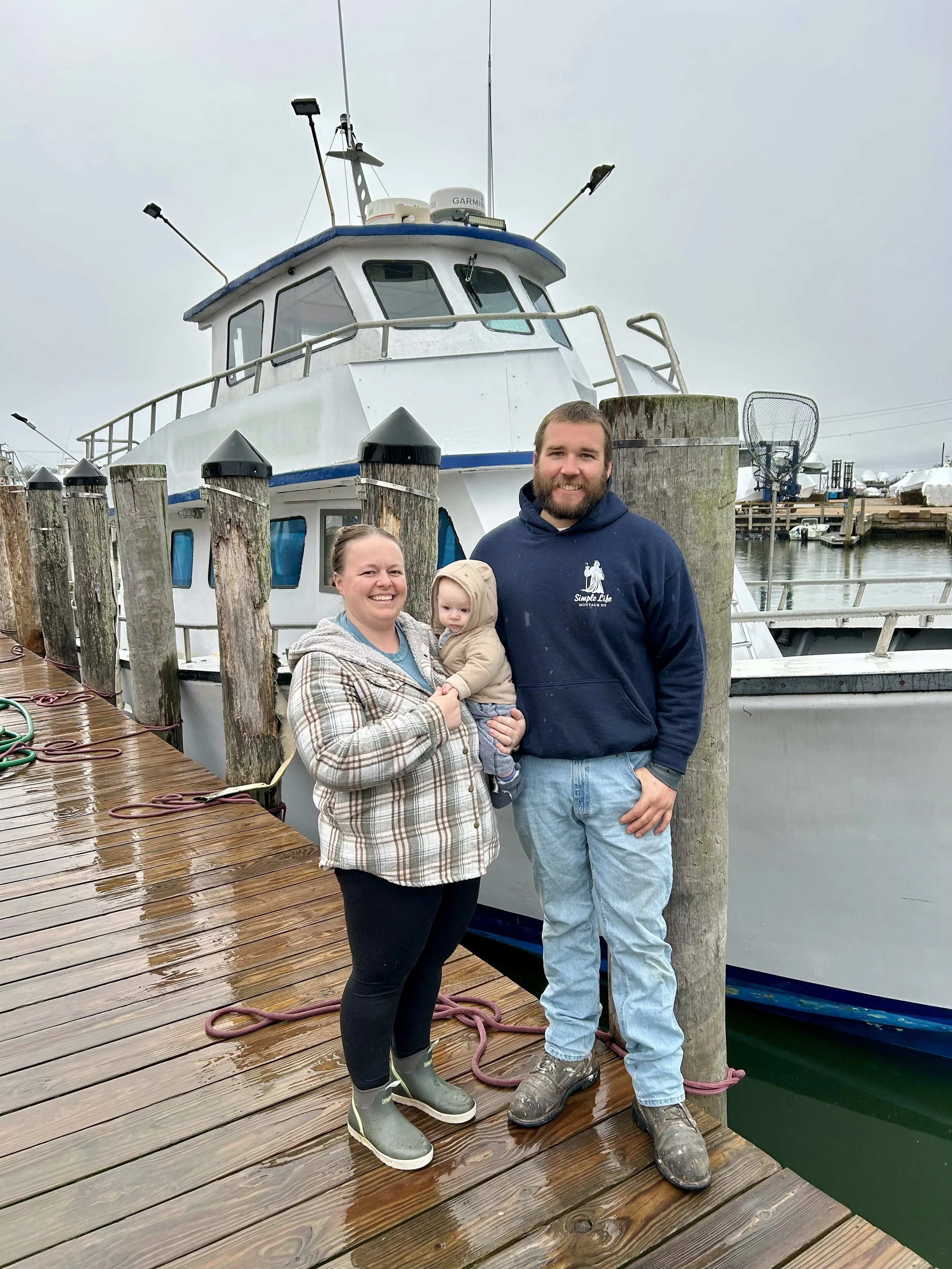 A smiling woman and man holding a baby standing on a wet wooden dock in front of a large boat tied to the dock, with other boats and docks in the background.