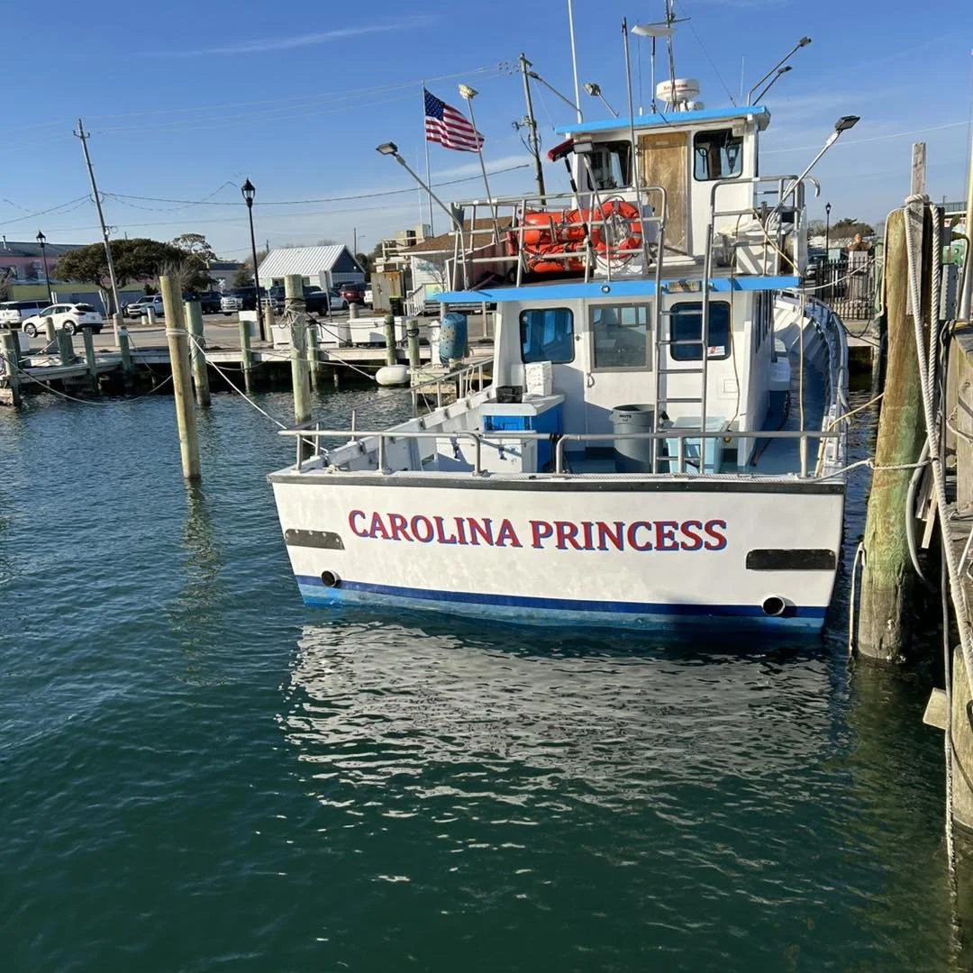 A boat named Carolina Princess docked at a marina, with an American flag flying above it, surrounded by colorful buildings and boats in the background.