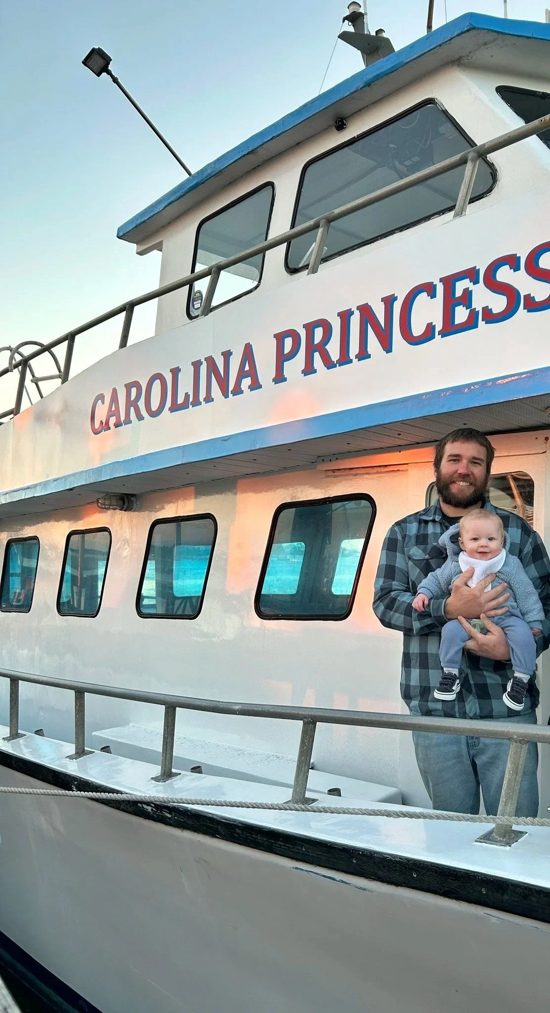 A man holding a baby on a boat named 'Carolina Princess' with a clear sky in the background.